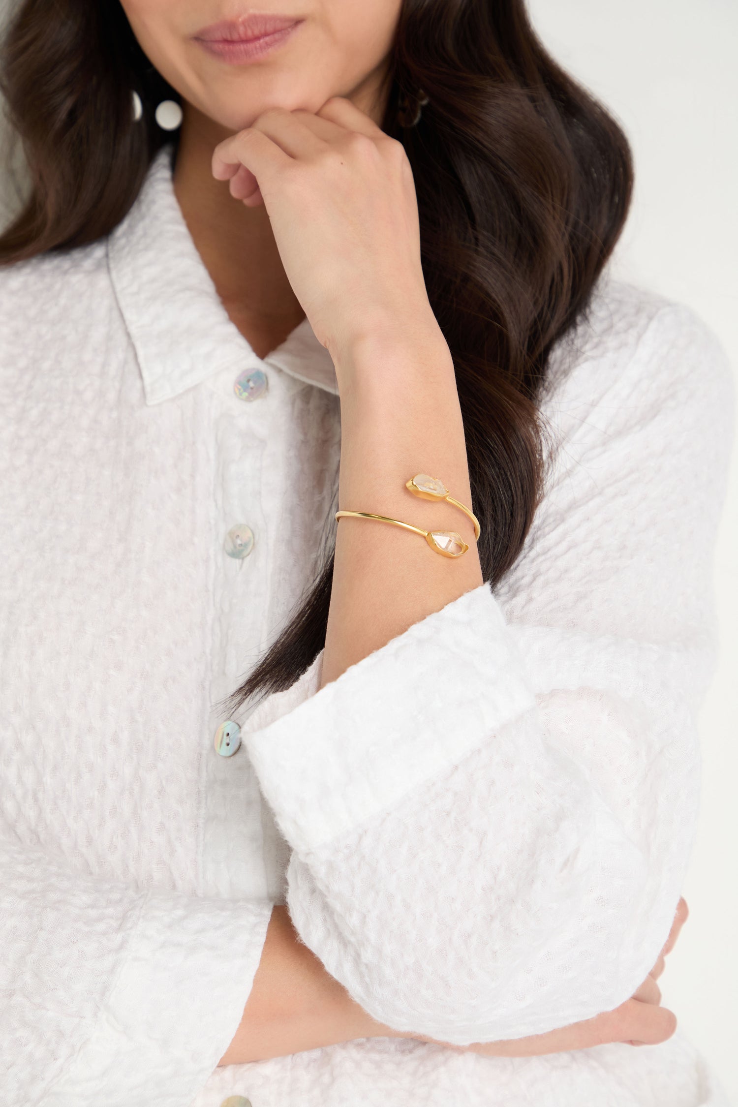 A woman in a textured white button-up shirt rests her chin on her hand, showcasing the Gold Plated Quartz Herkimer Diamond Bracelet with an open cuff design on her wrist.