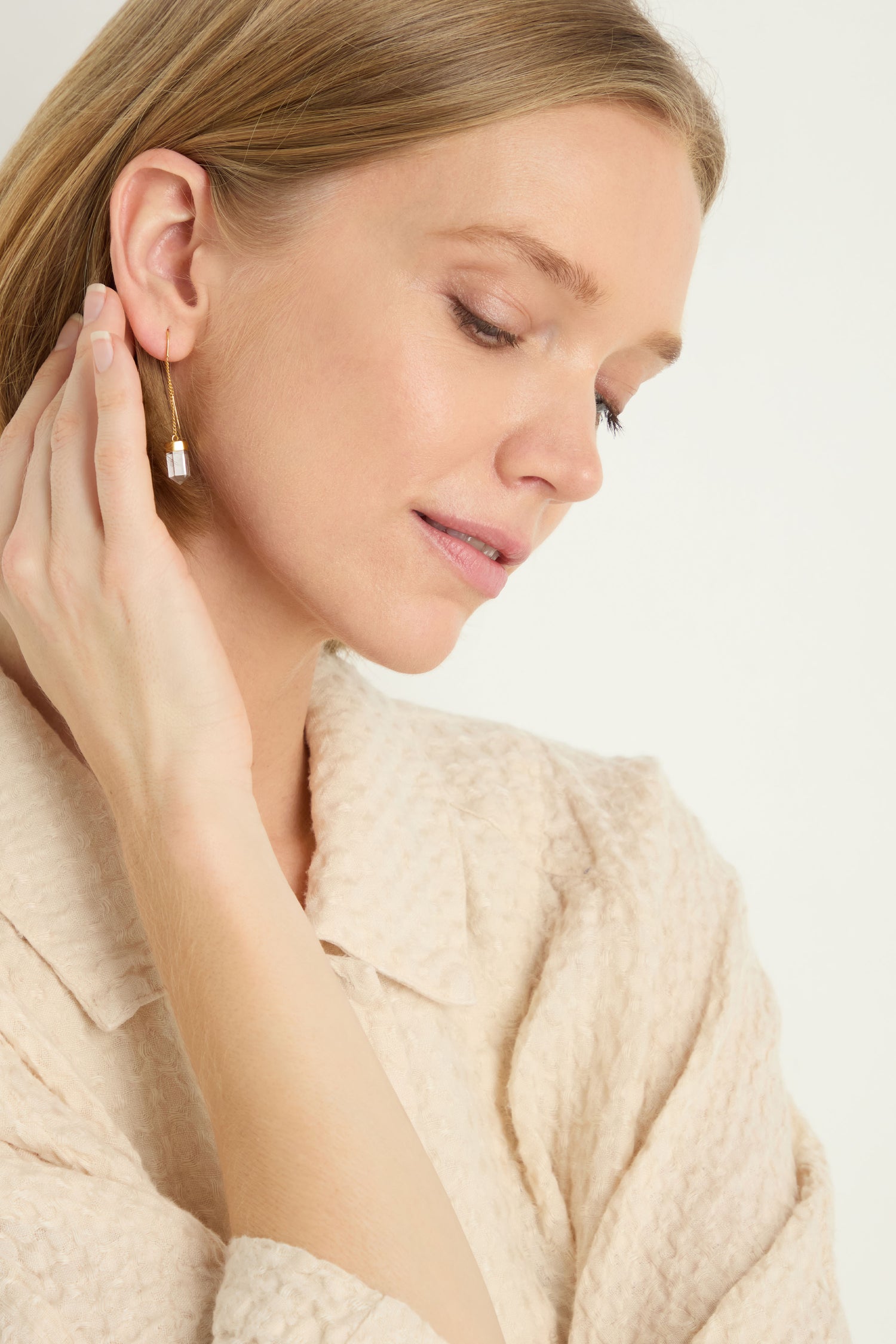 A woman in a textured beige top touches her ear and looks down, showcasing Hand Crafted Clear Quartz Crystal Drop Earrings.