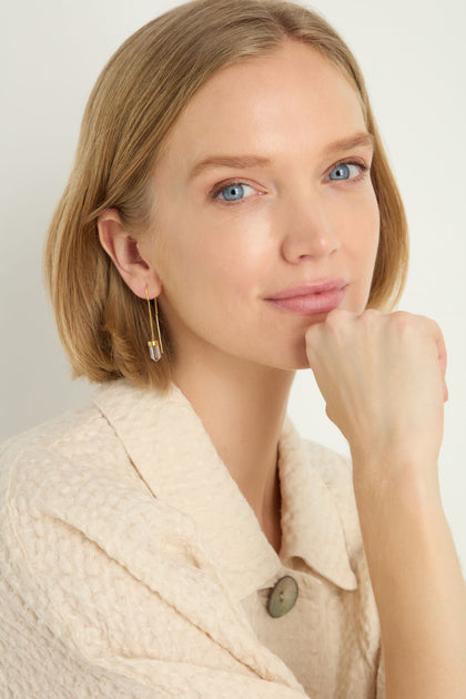 A woman with short blonde hair and blue eyes, wearing a textured beige jacket and Hand Crafted Clear Quartz Crystal Drop Earrings, rests her chin on her hand and looks at the camera.