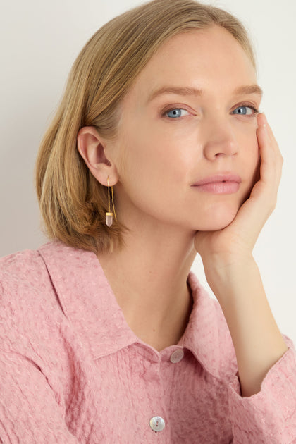 Woman with blonde bob wears a textured pink blouse and Rose Quartz Crystal Drop Earrings, resting her face on her hand and looking at the camera with a neutral expression.