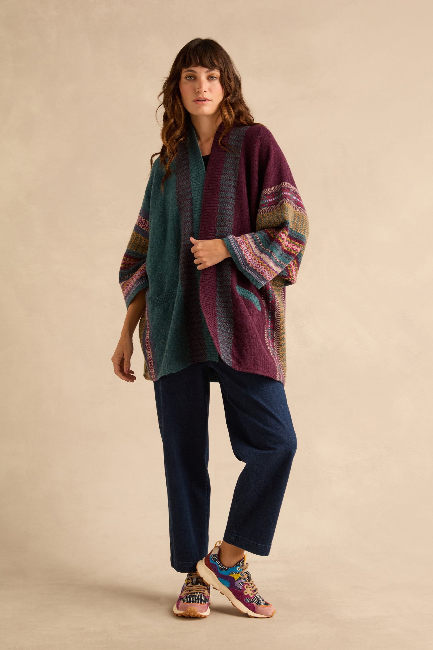A woman wearing an open, color-blocked Hunter Montrose Blanket Cardigan with patterned sleeves over dark jeans and colorful sneakers.