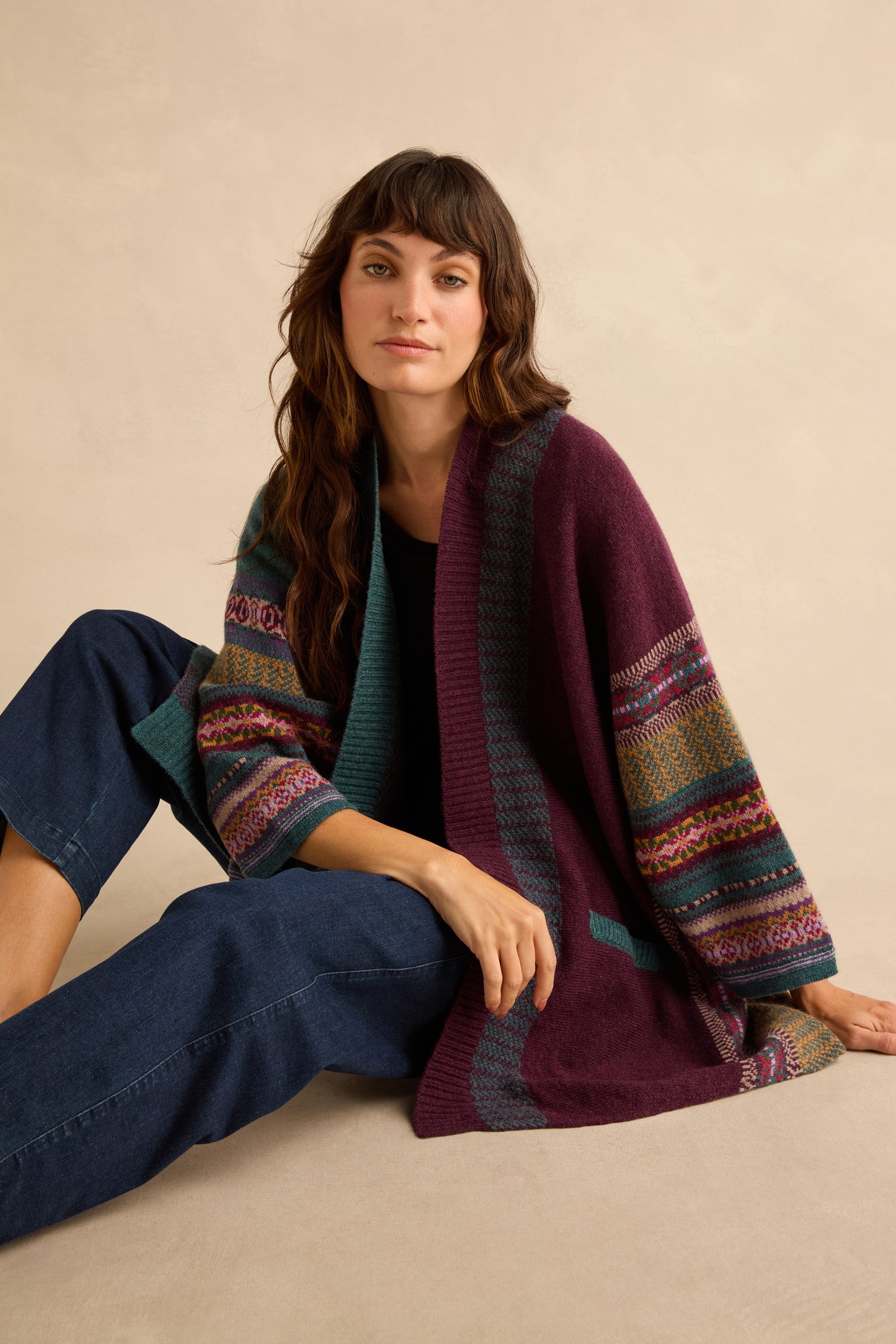A woman sits on the floor wearing a multi-colored geometric Fair Isle Hunter Montrose Blanket Cardigan paired with dark blue jeans.