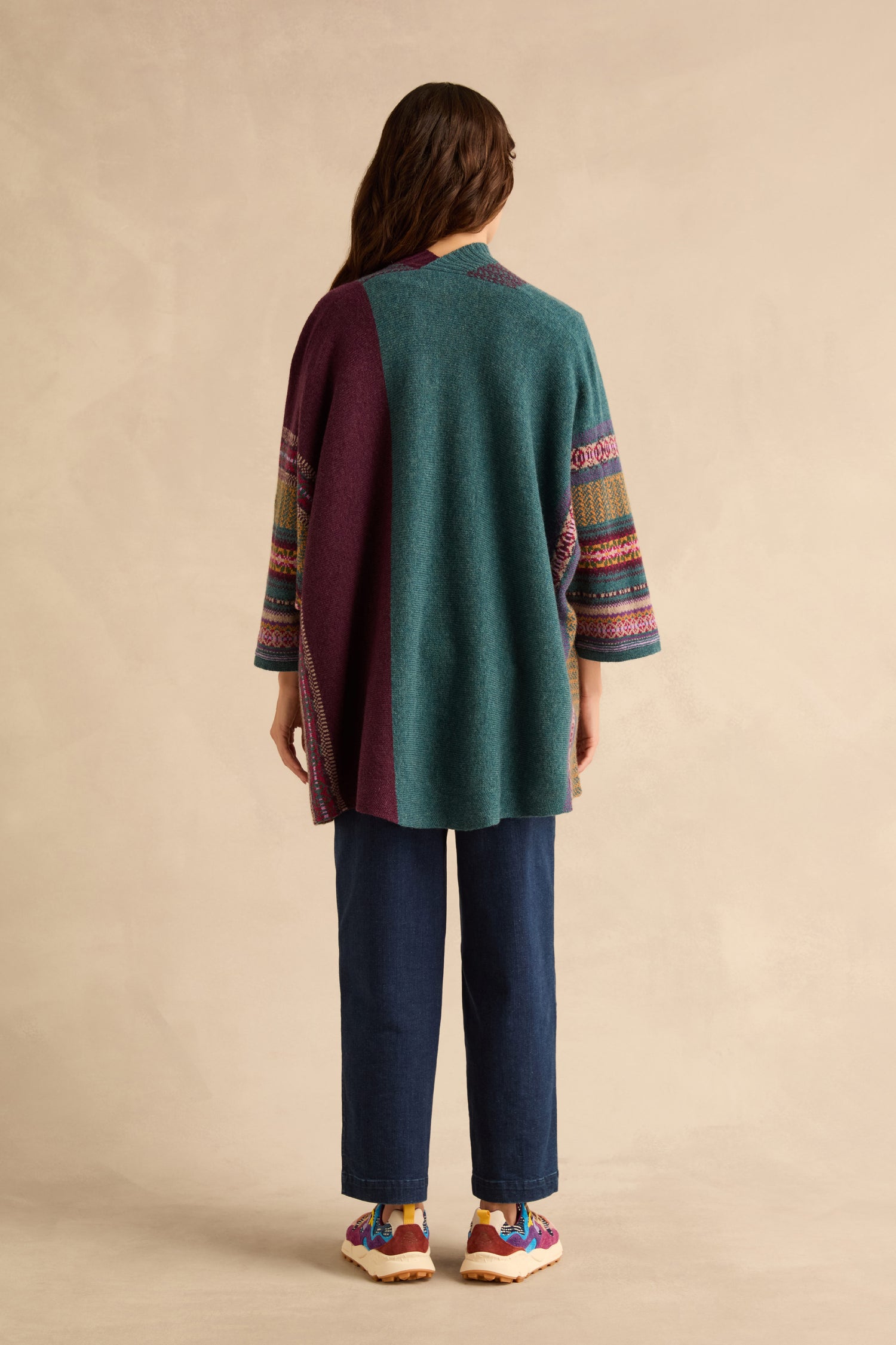 A person seen from behind wears a Hunter Montrose Blanket Cardigan with a Fair Isle pattern, three-quarter length sleeves, dark blue jeans, and colorful sneakers.
