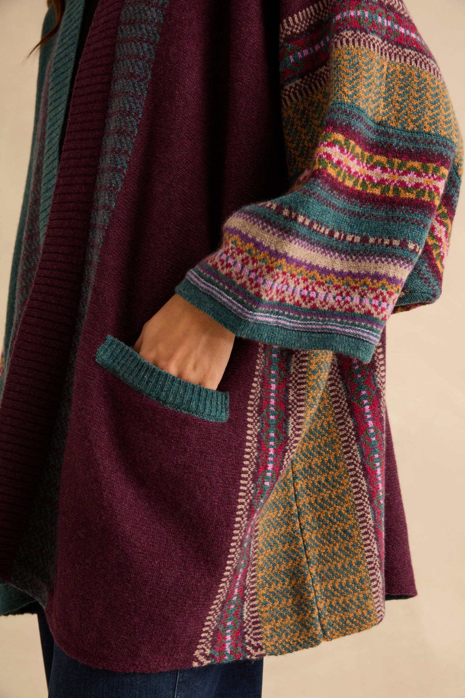 A person wearing a dark purple Hunter Montrose Blanket Cardigan with teal trim and Fair Isle patterned sleeves, shown with their hand in a patch pocket.