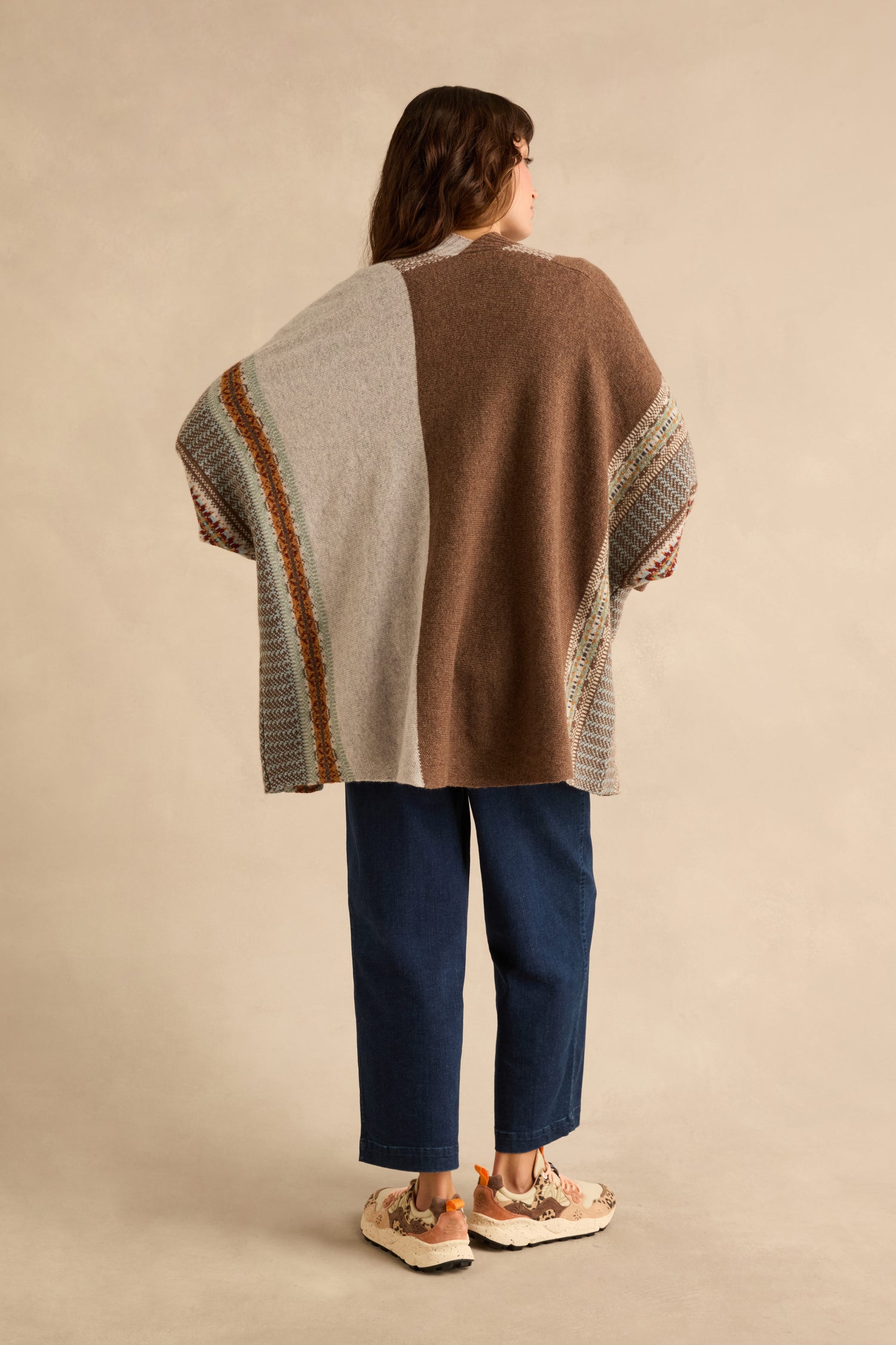 A person seen from the back wearing an oversized Mushroom Montrose Blanket Cardigan in brown and beige with a Fair Isle pattern, dark blue jeans, and patterned sneakers.