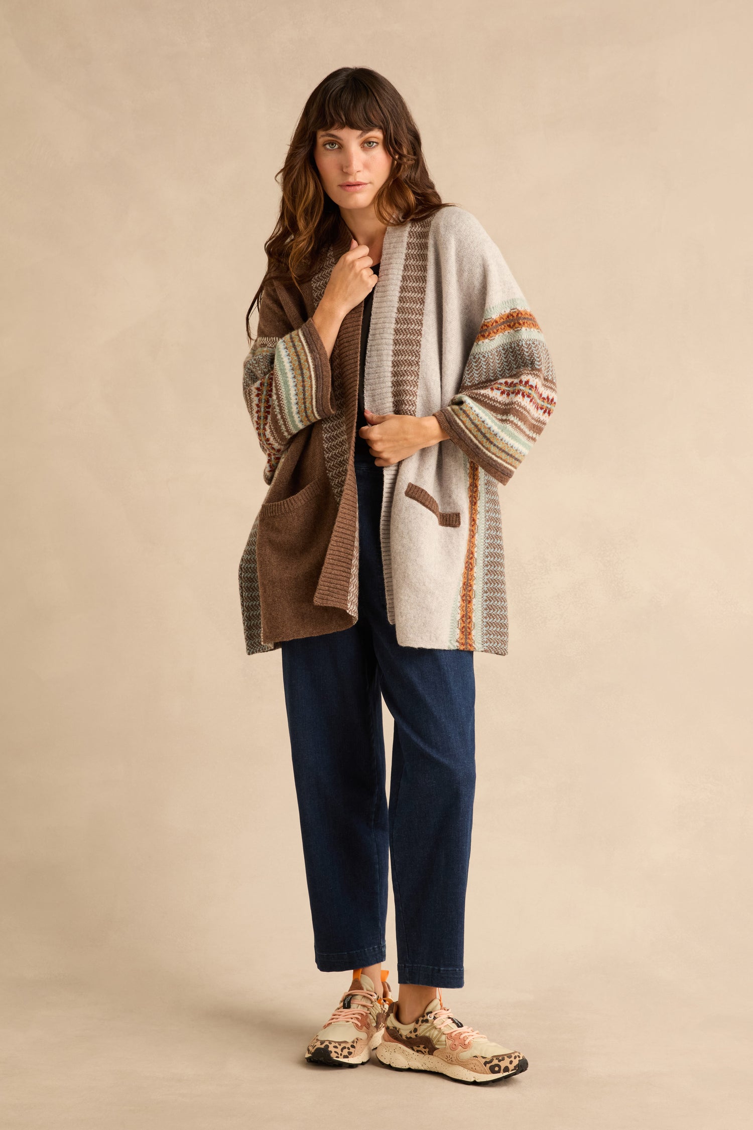 A person wearing a Mushroom Montrose Blanket Cardigan with a Fair Isle pattern over a dark shirt and blue jeans, paired with patterned sneakers.