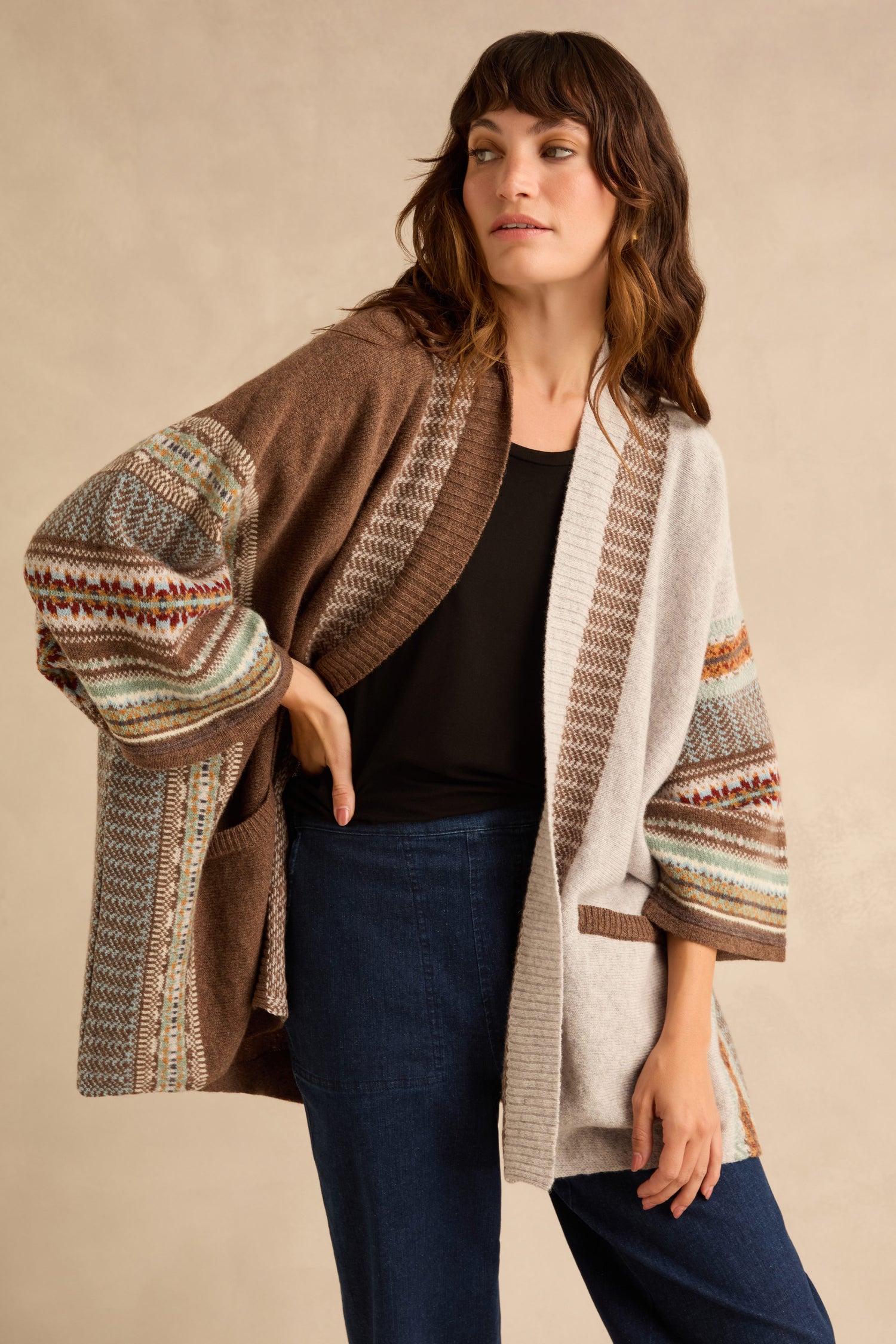 A person wears an open Mushroom Montrose Blanket Cardigan, featuring brown and beige color-blocked panels with Fair Isle patterned sleeves, over a black top and dark blue jeans.