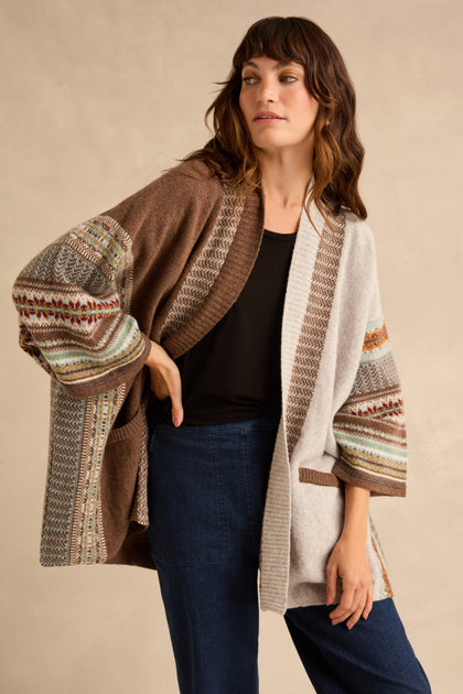 A person wears an open Mushroom Montrose Blanket Cardigan, featuring brown and beige color-blocked panels with Fair Isle patterned sleeves, over a black top and dark blue jeans.