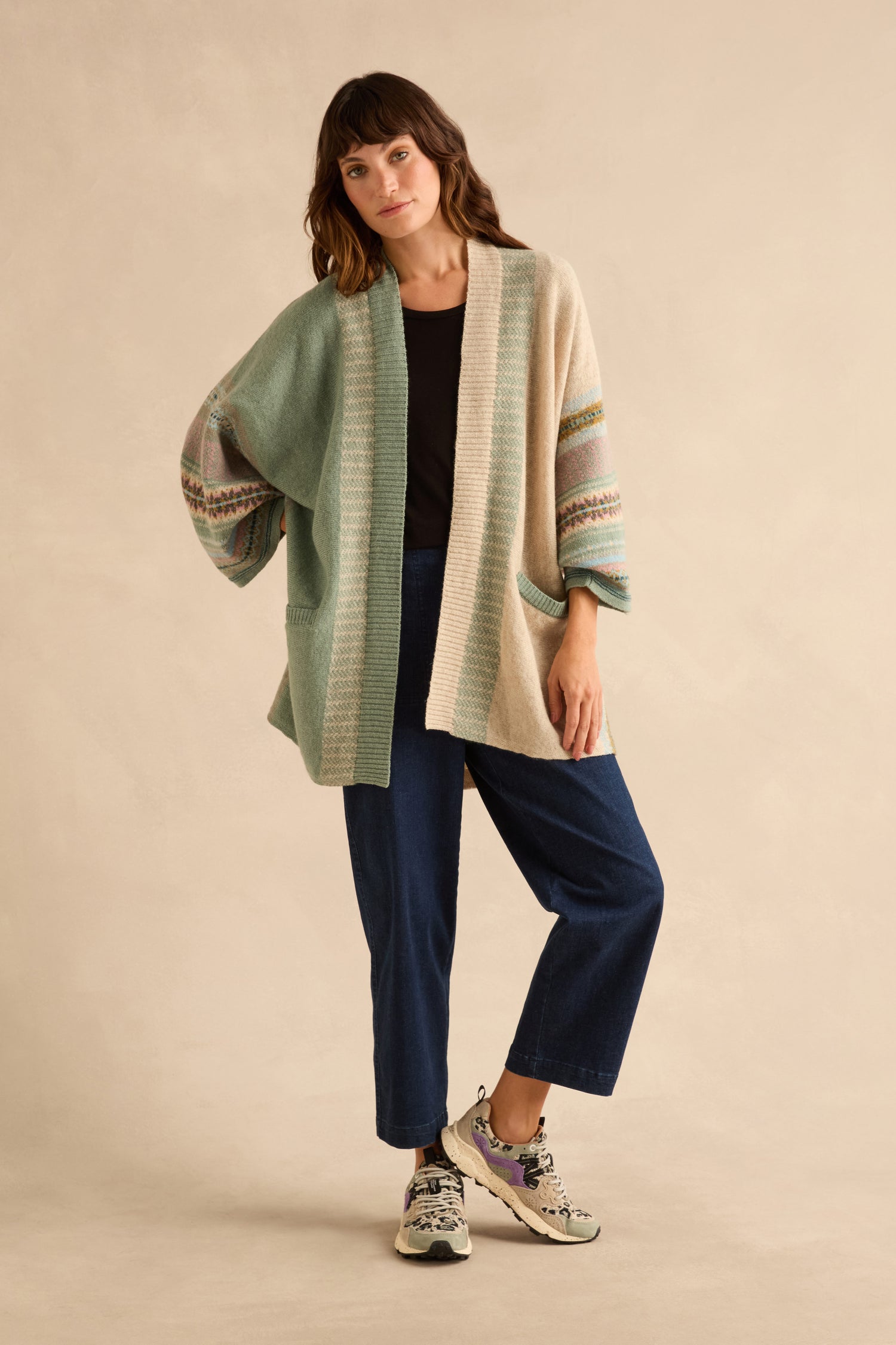 A woman wears an open, color-blocked Sage Montrose Blanket Cardigan in sage green and beige with patterned trim over a black top and dark jeans.