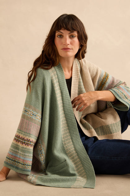 A person wears an open, sage green and beige Sage Montrose Blanket Cardigan with wide sleeves over a black top and blue jeans.