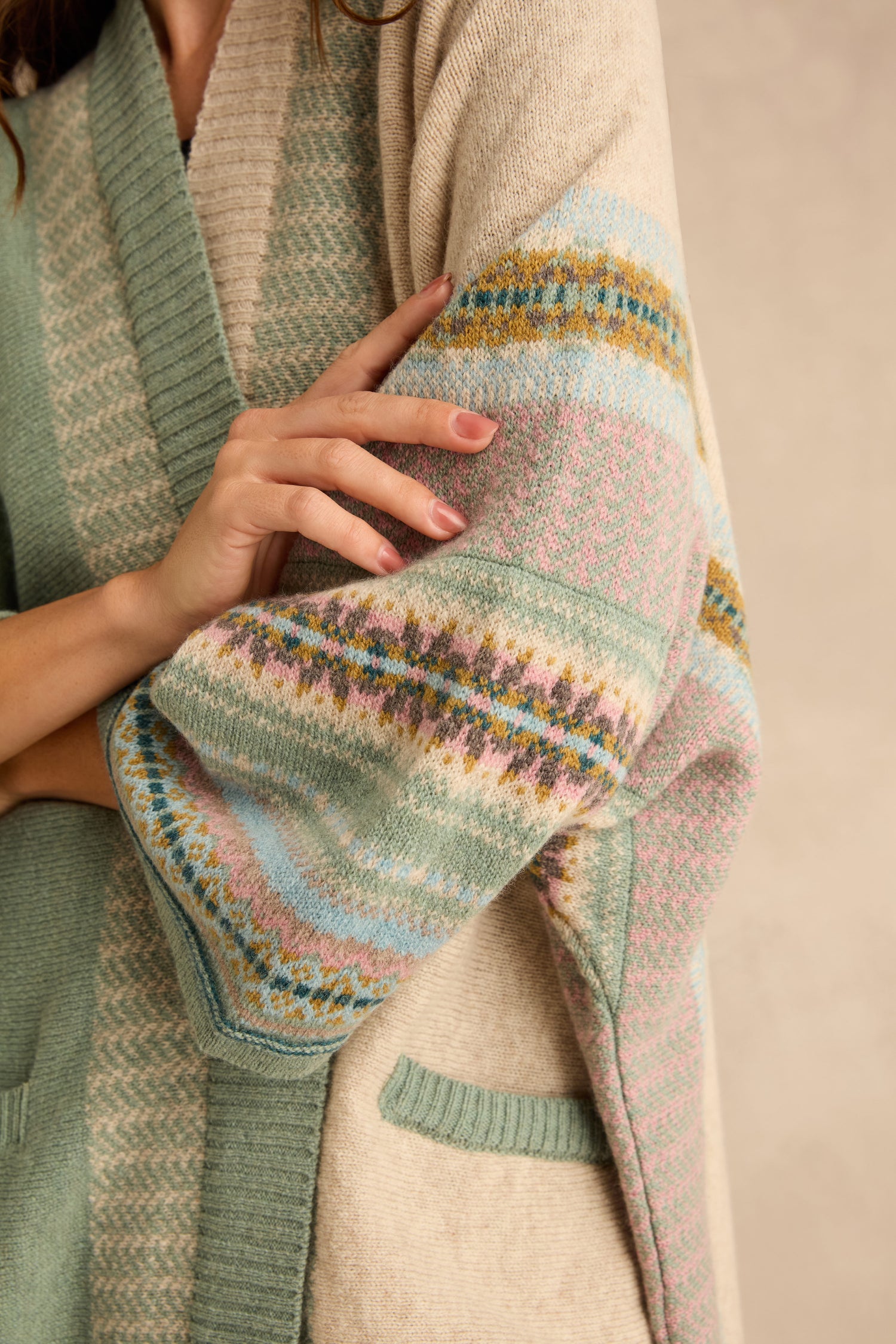 A person wearing a sage green and cream-colored Sage Montrose Blanket Cardigan in merino lambswool, featuring an open Fair Isle pattern and wide sleeves.
