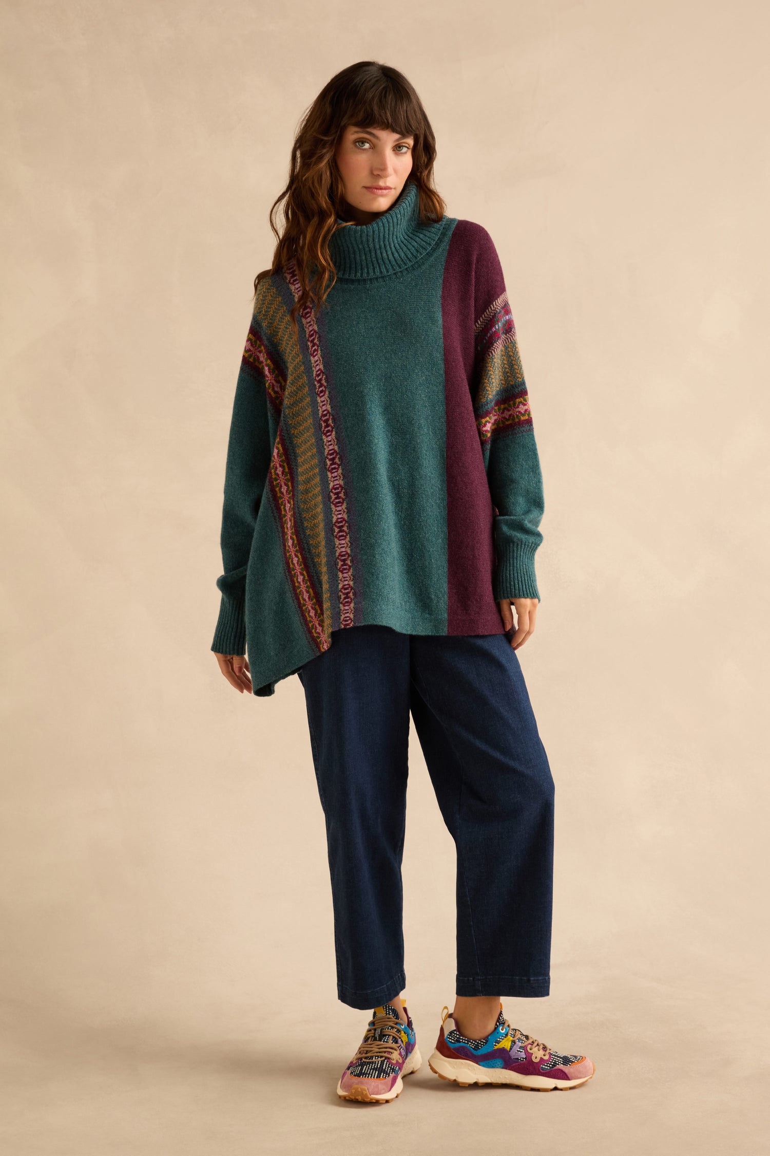 A person wears an oversized, multicolored Hunter Montrose Cowl Neck Sweater with a geometric pattern, dark blue jeans, and colorful athletic sneakers.