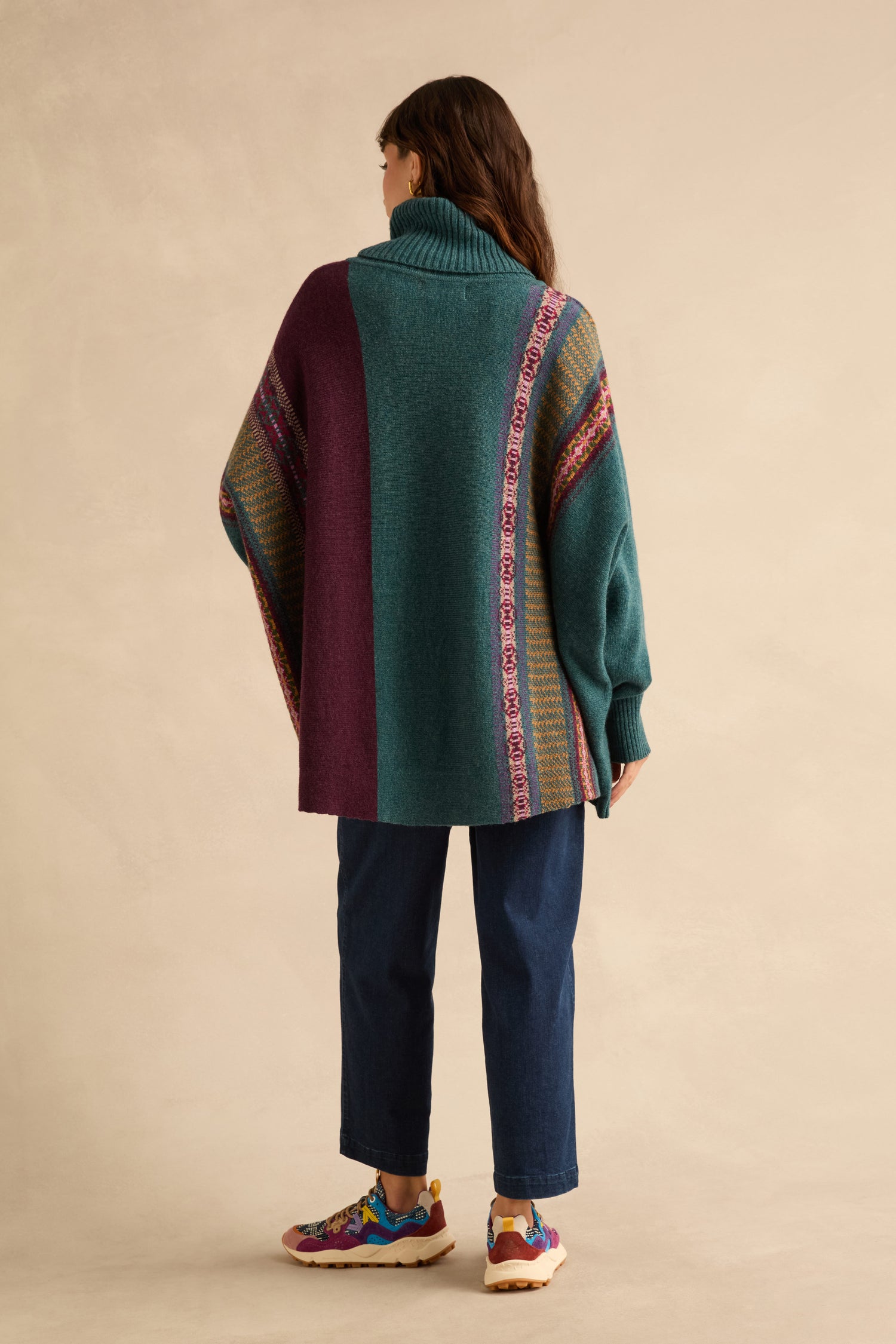 A person wears a Hunter Montrose Cowl Neck Sweater featuring geometric panels in teal and purple, styled with dark blue jeans and colorful sneakers.