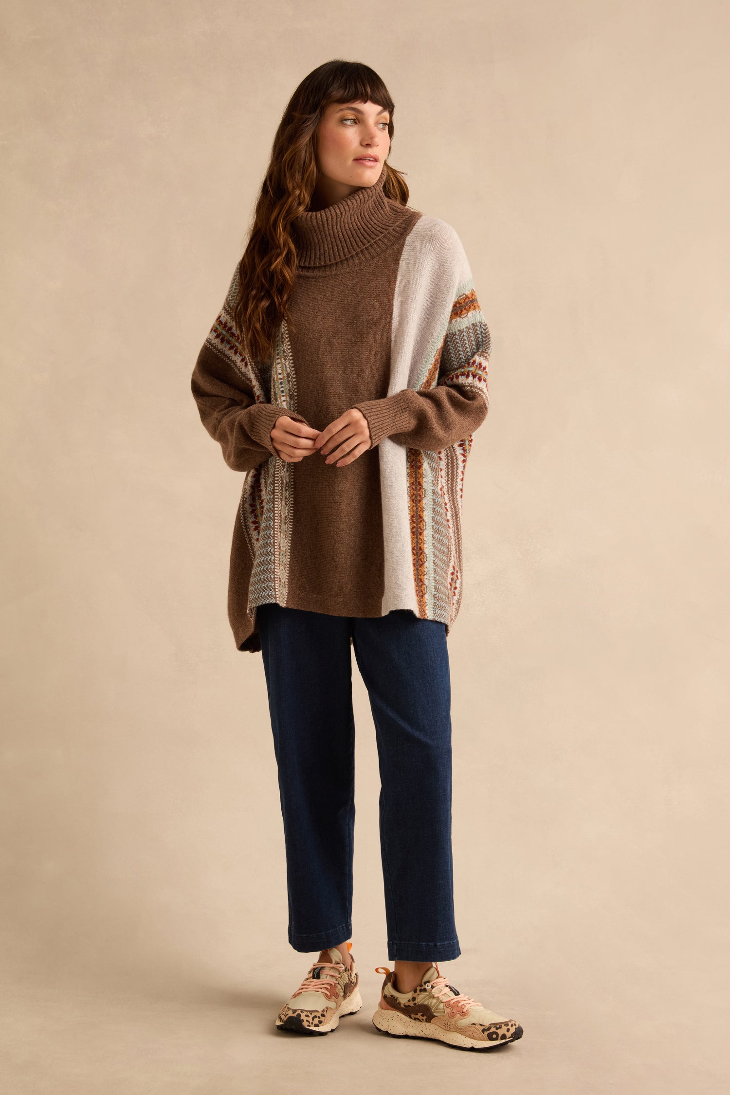 A person wearing a Mushroom Montrose Cowl Neck Sweater in brown and cream color-blocked merino lambswool, paired with dark blue jeans and patterned sneakers.