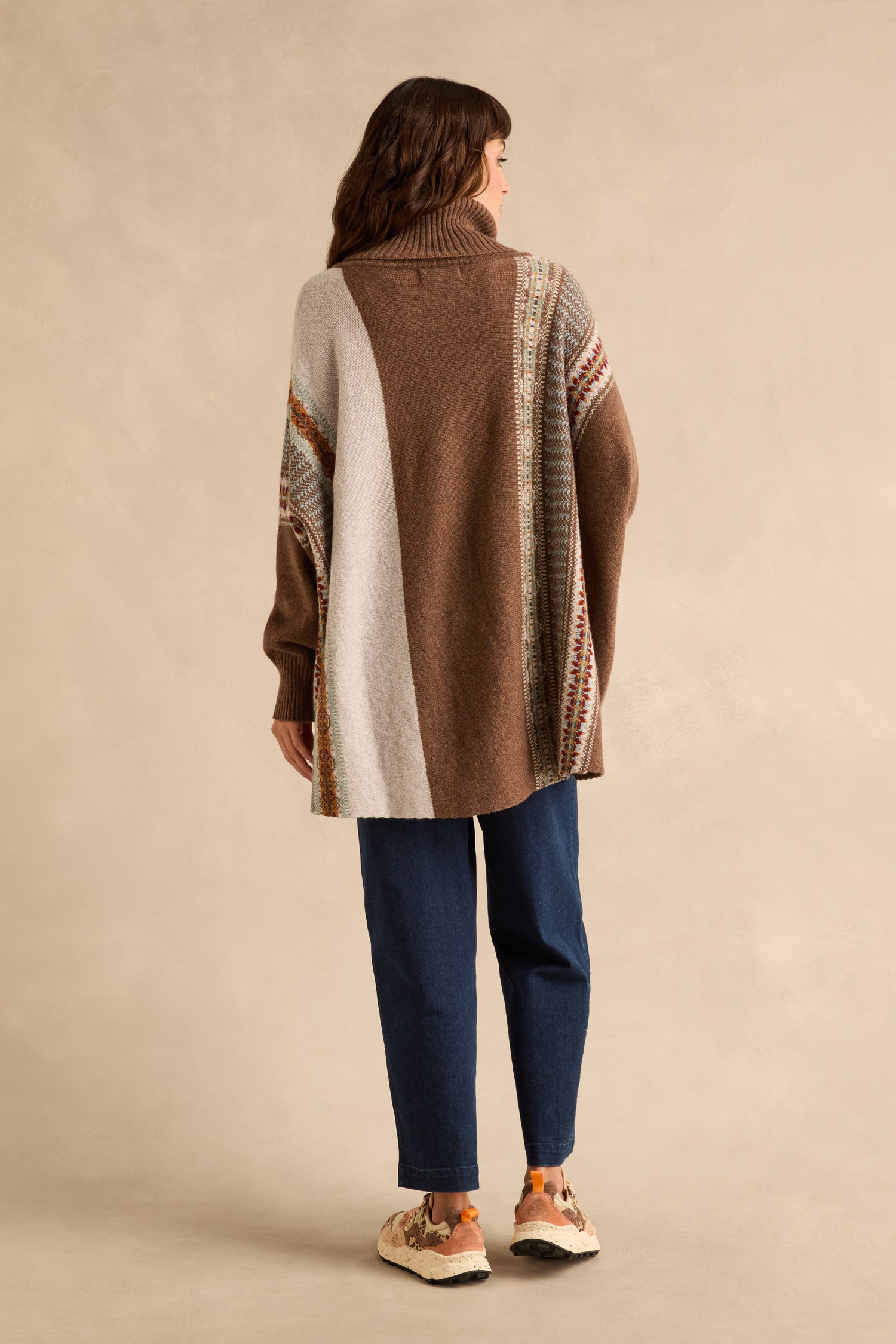 A person seen from behind wears an oversized Mushroom Montrose Cowl Neck Sweater with color-blocked panels and patterned trim, paired with dark blue jeans and athletic shoes.