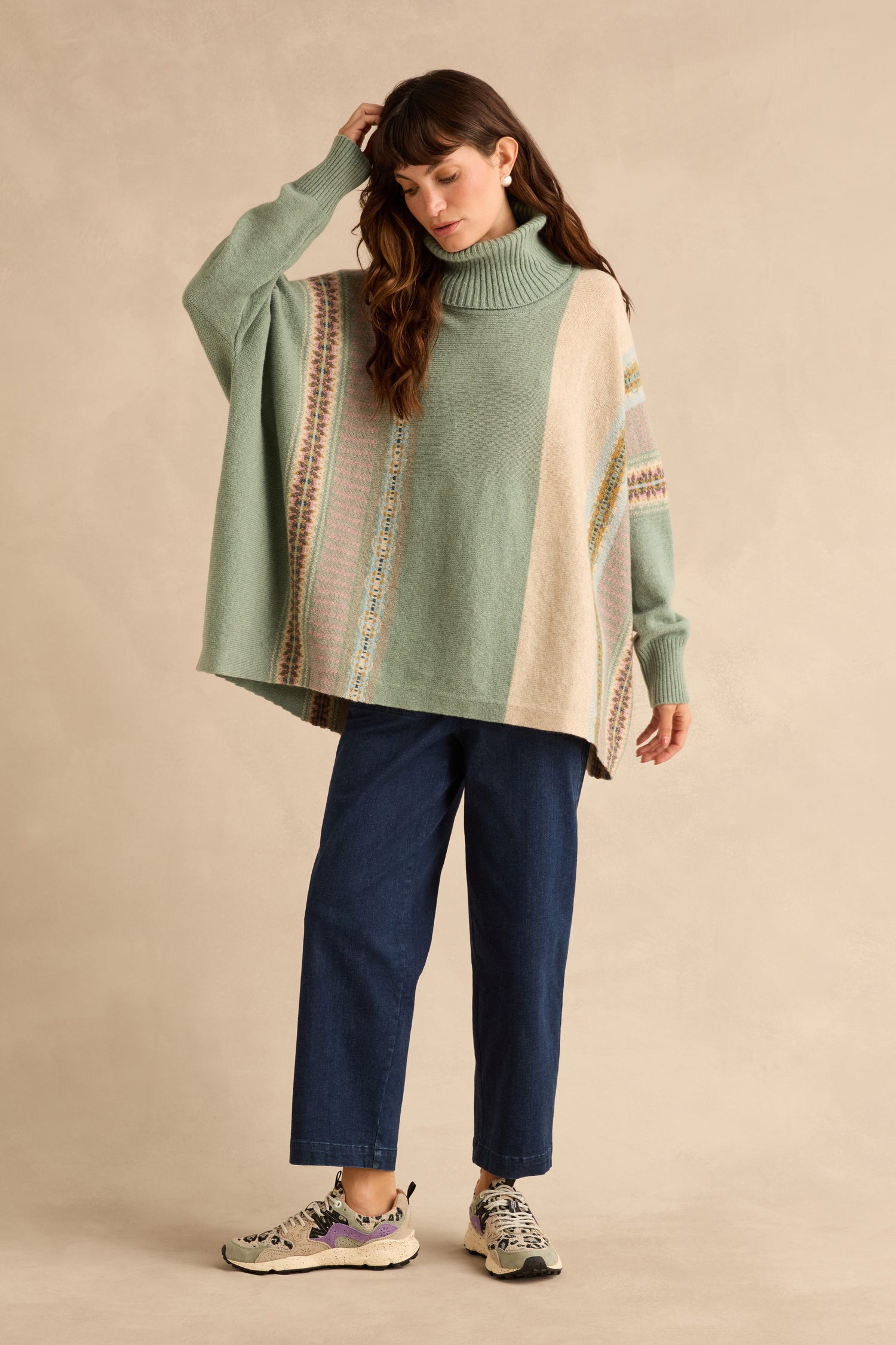 A person wears a sage green Sage Montrose Cowl Neck Sweater featuring vertical patterned panels, dark blue jeans, and patterned sneakers.