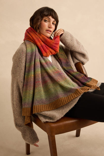 A person wears a Multicolour Treeline Merino Wool Scarf, a long, kid mohair blend ribbed knit accessory with a red, orange, green, and brown gradient and solid brown ribbed trim at the ends.