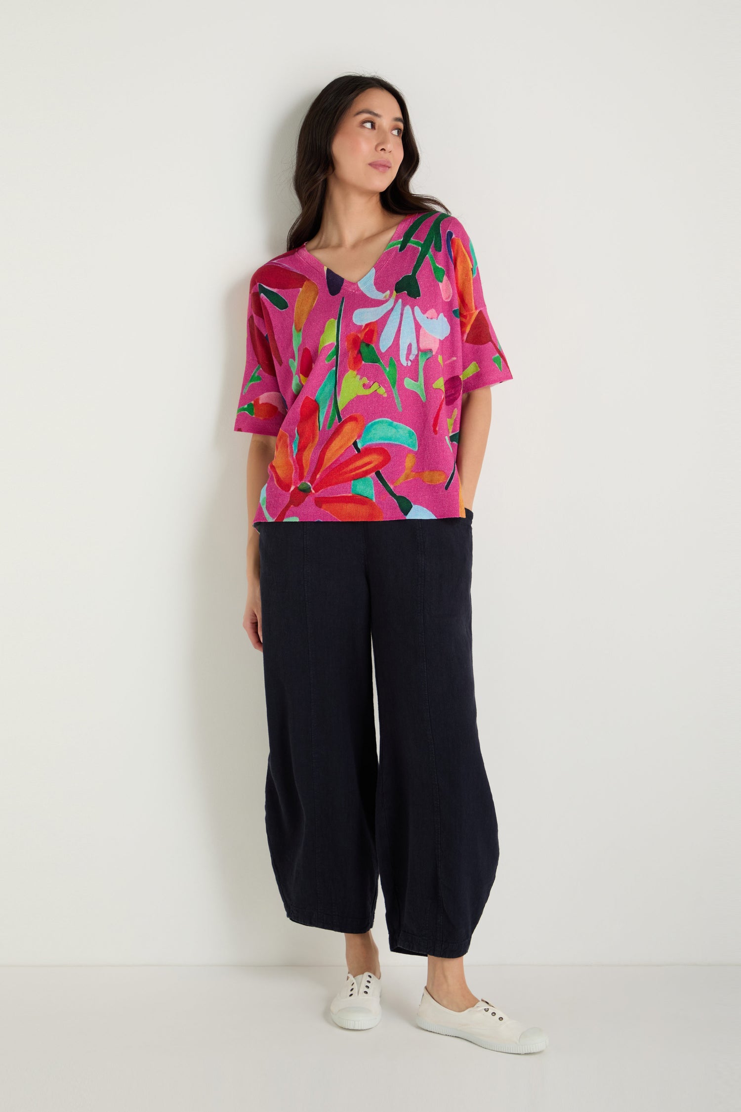 A woman stands against a white wall wearing the Abstract Floral V-Neck Knit, paired with loose black pants and white sneakers. She looks to her left with her hands in her pockets.