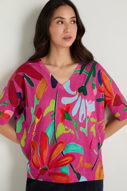 Woman models the Abstract Floral V-Neck Knit sweater featuring a pink base with red, orange, green, and white abstract floral patterns, posed against a simple light background.