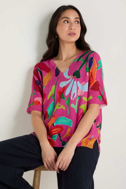 A woman sits on a stool wearing the Abstract Floral V-Neck Knit and dark pants, looking to the side against a plain light background.