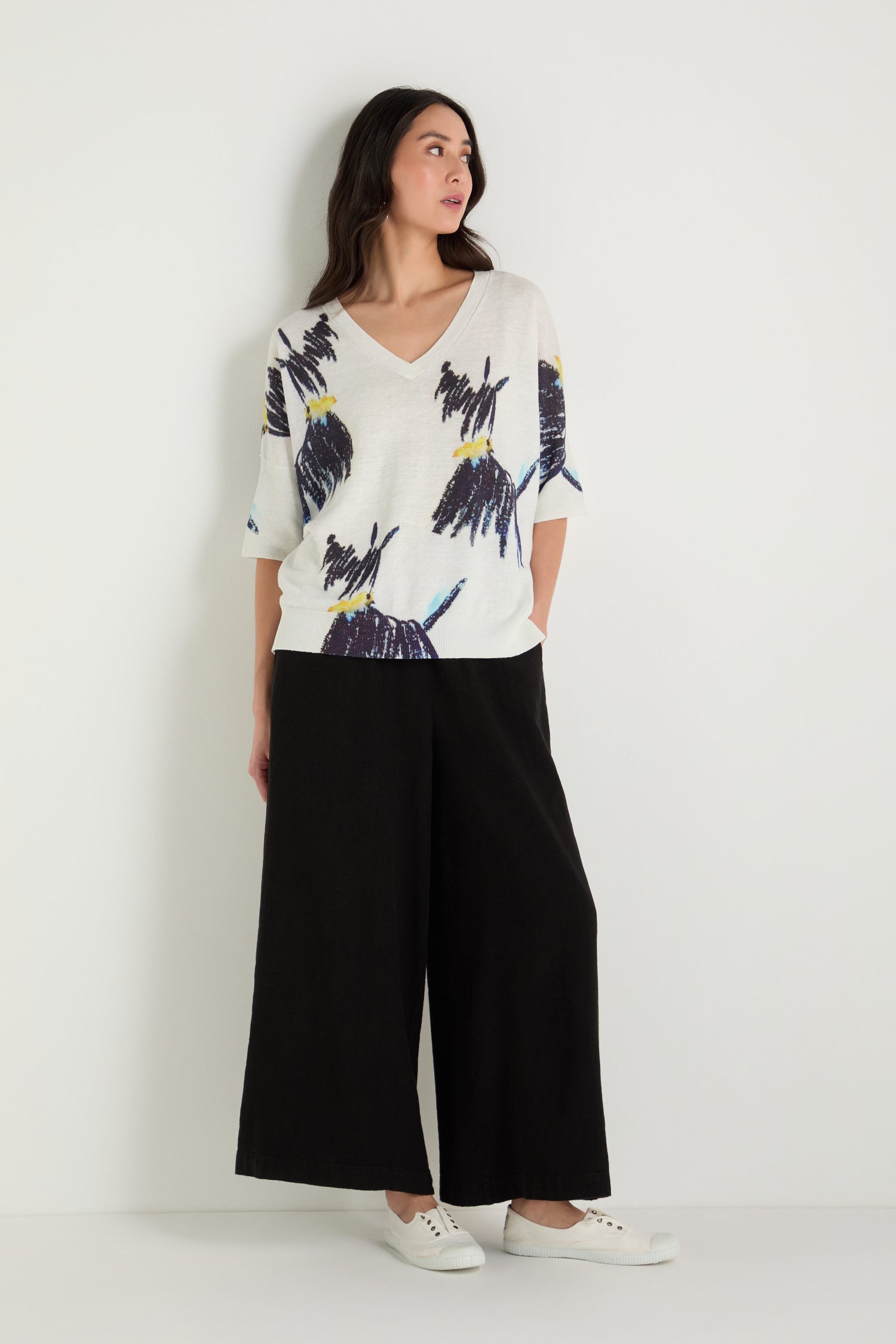 A woman stands against a white wall wearing the Scottie Linen Cotton V-Neck Knit with an abstract Scottie print, paired with black wide-leg pants and white sneakers.