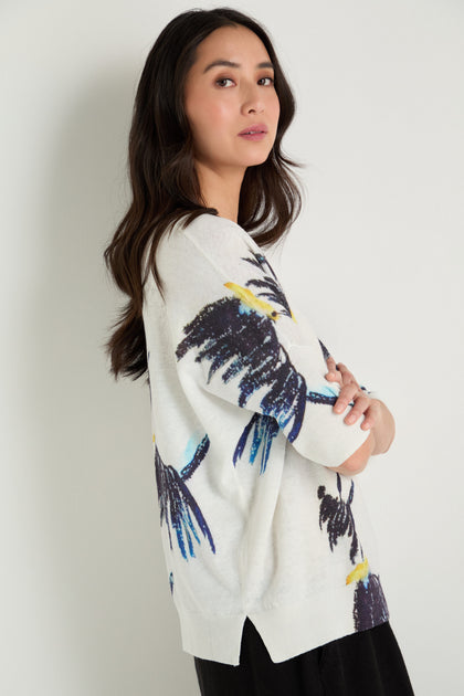 Woman with long dark hair stands sideways against a white wall, wearing the Scottie Linen Cotton V-Neck Knit featuring a colorful bird print and black pants.