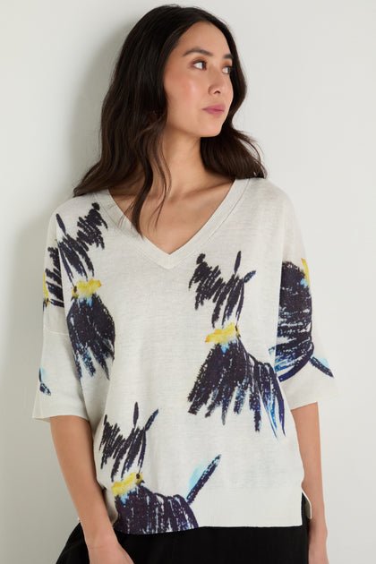 A woman with long dark hair wears the Scottie Linen Cotton V-Neck Knit, a white sweater featuring a bold abstract floral print in dark purple, blue, and yellow.