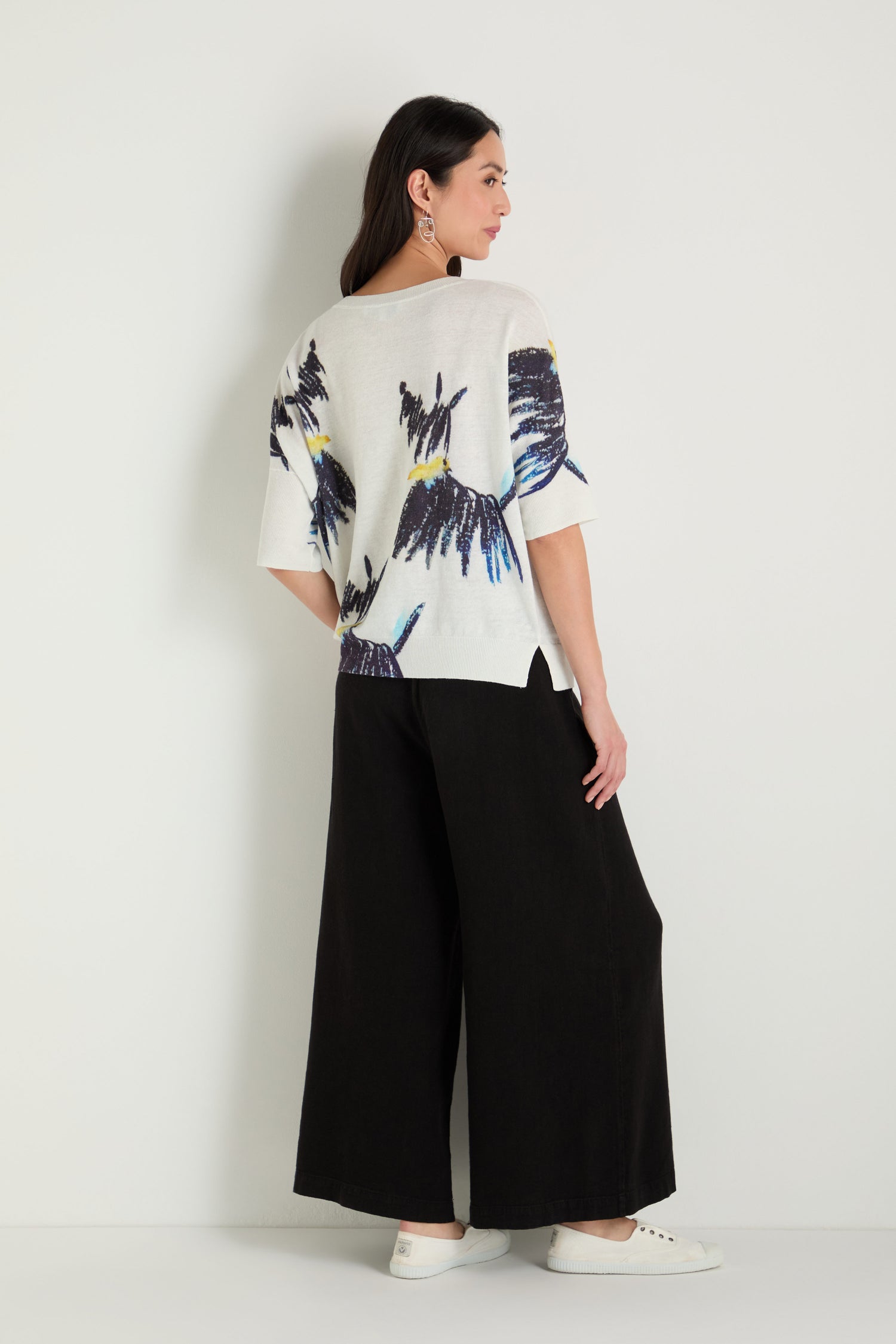 A woman stands sideways against a white wall, wearing the Scottie Linen Cotton V-Neck Knit sweater with a large bird print on the back, paired with wide-leg black pants and white shoes.