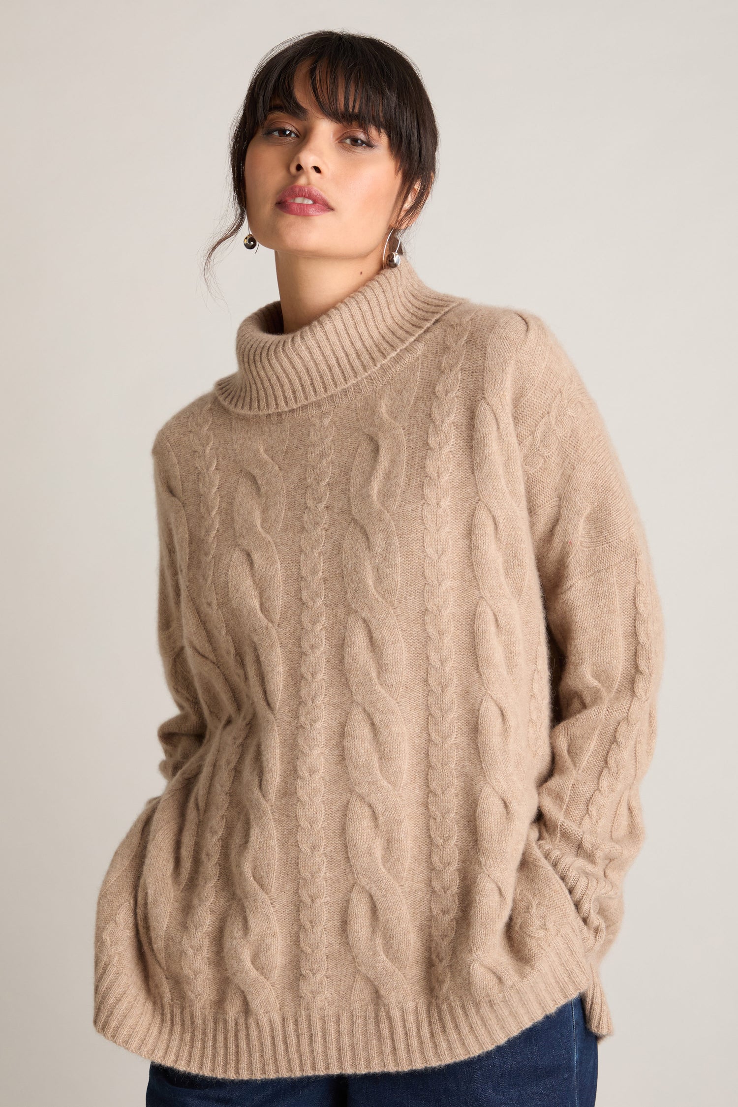 Person wearing a Loose Cable Cashmere Knit turtleneck sweater in beige with hands in pockets.