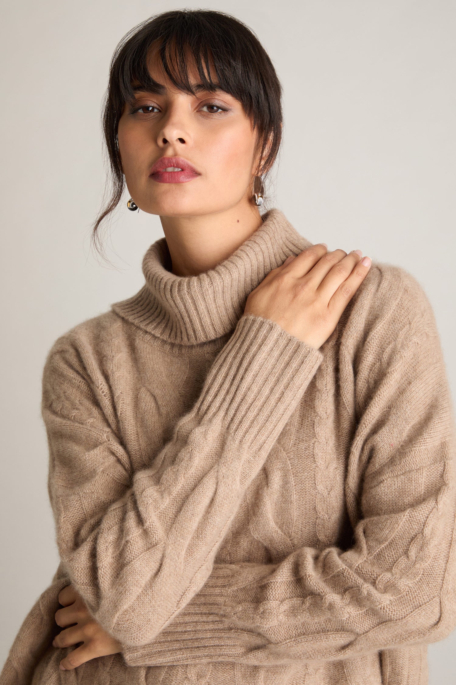 A person with dark hair rests a hand on their shoulder while wearing a beige Loose Cable Cashmere Knit sweater.