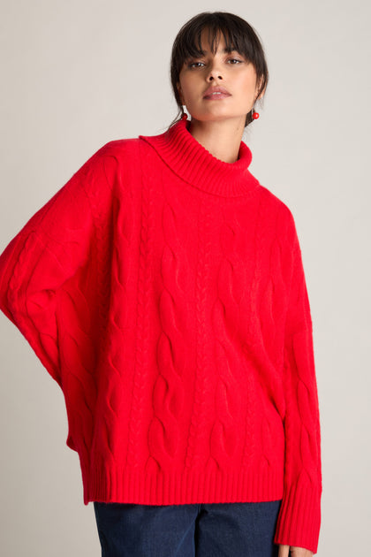 A person is wearing a bright red, Loose Cable Cashmere Knit turtleneck sweater from Estheme, posing with one hand in their pocket against a plain background.