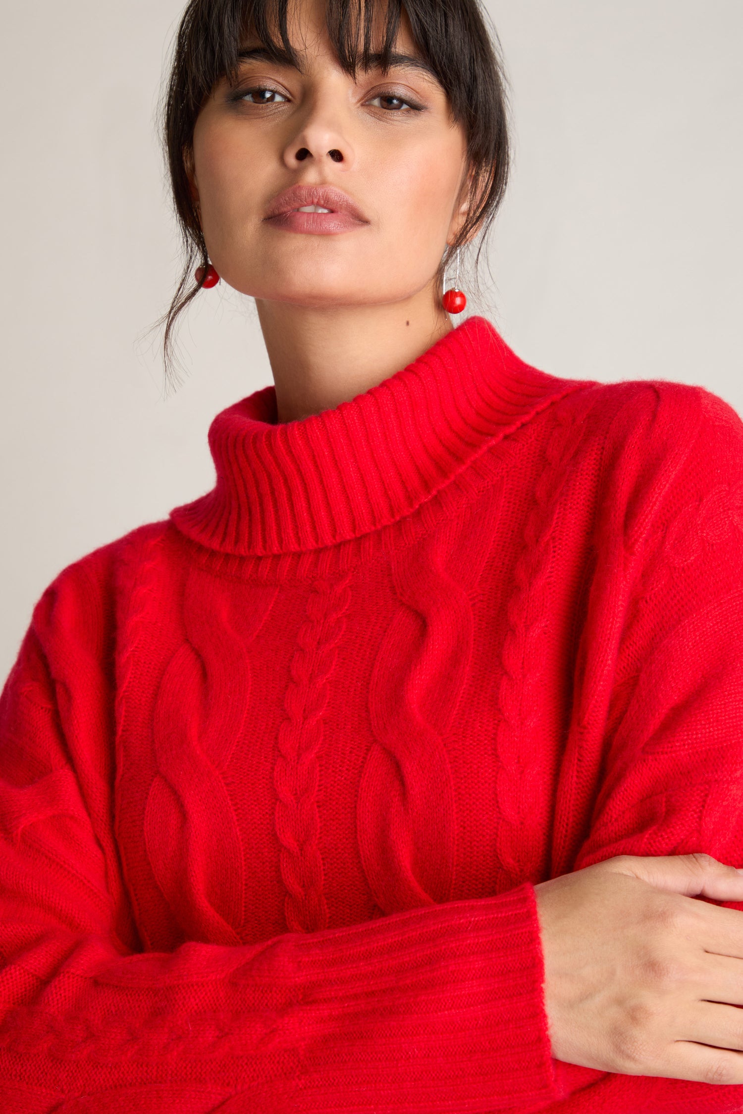 A person stands wearing a bright red Loose Cable Cashmere Knit sweater and red earrings, with a neutral expression, looking slightly off-camera.