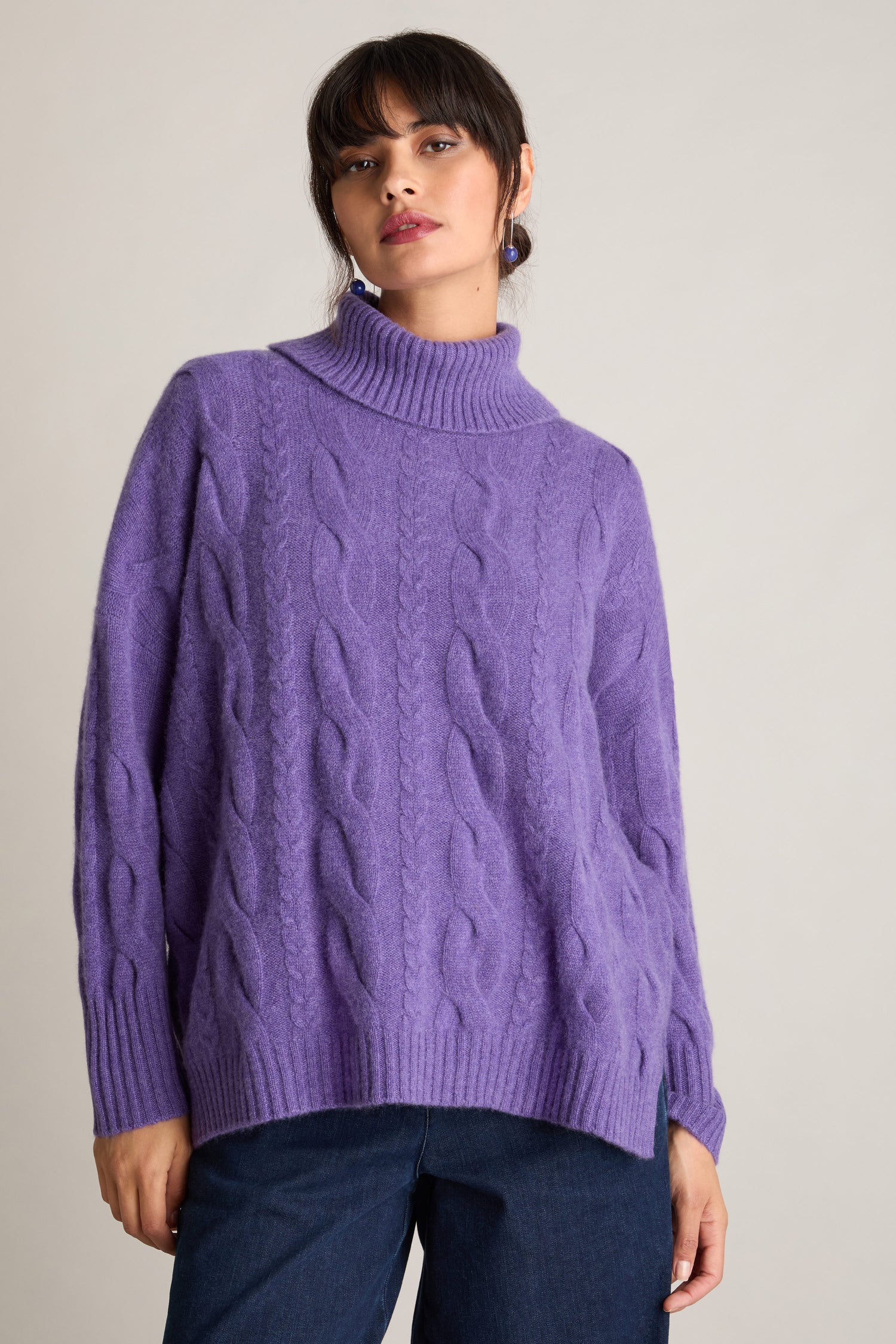 A person wearing a purple Loose Cable Cashmere Knit turtleneck sweater and blue jeans stands against a plain background.