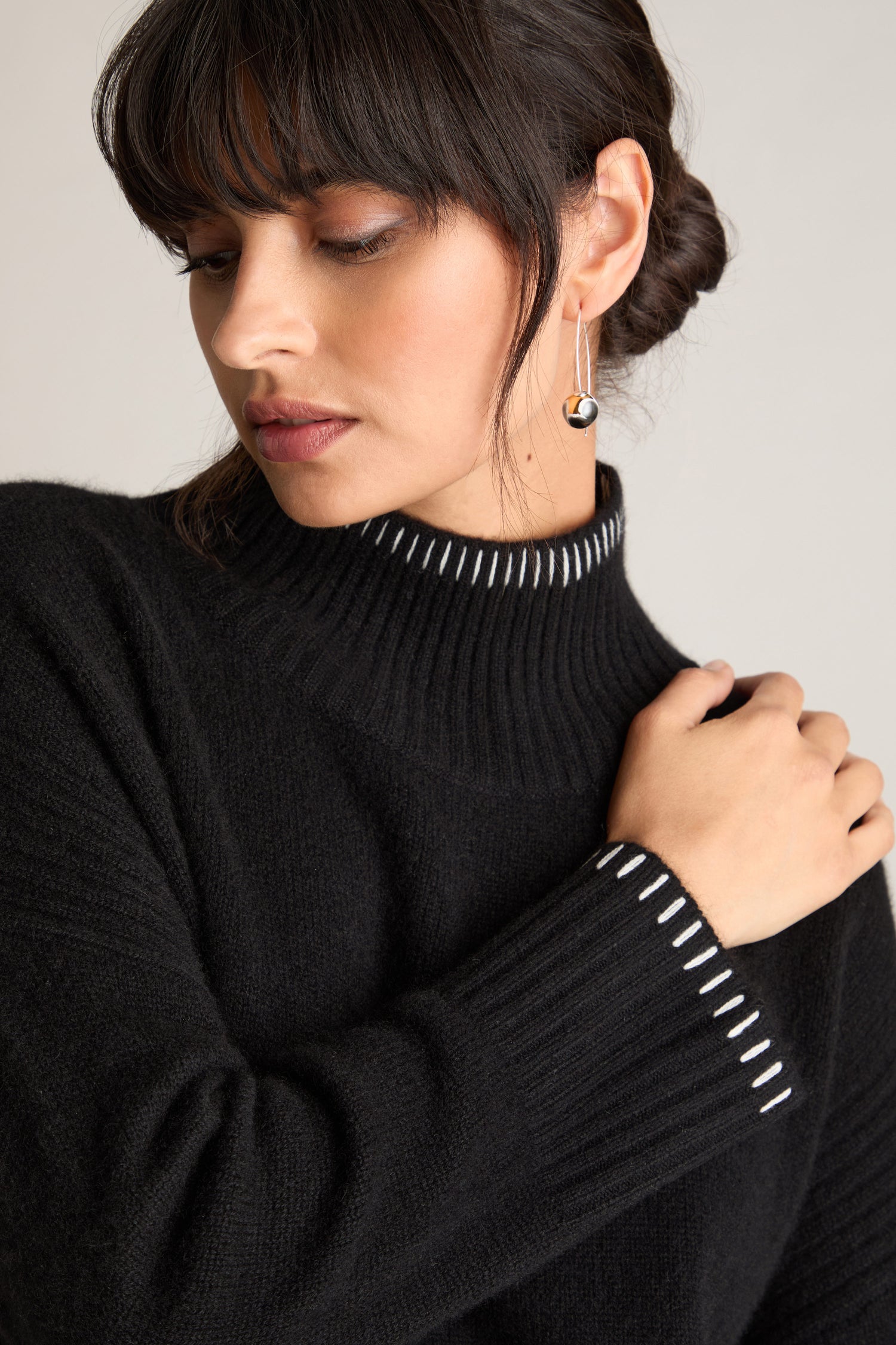 A woman with dark hair and bangs wears an Estheme Blanket Stitch Funnel Neck Cashmere Knit sweater and a pair of drop earrings, posing with her right hand on her left shoulder.