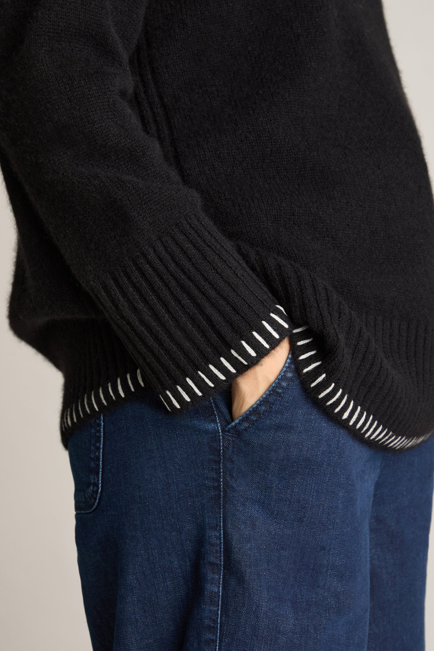 Side view of a person wearing the Blanket Stitch Funnel Neck Cashmere Knit in black with white stitches on the cuffs and hem, paired with blue jeans. The person's left hand is in the pocket of the jeans.