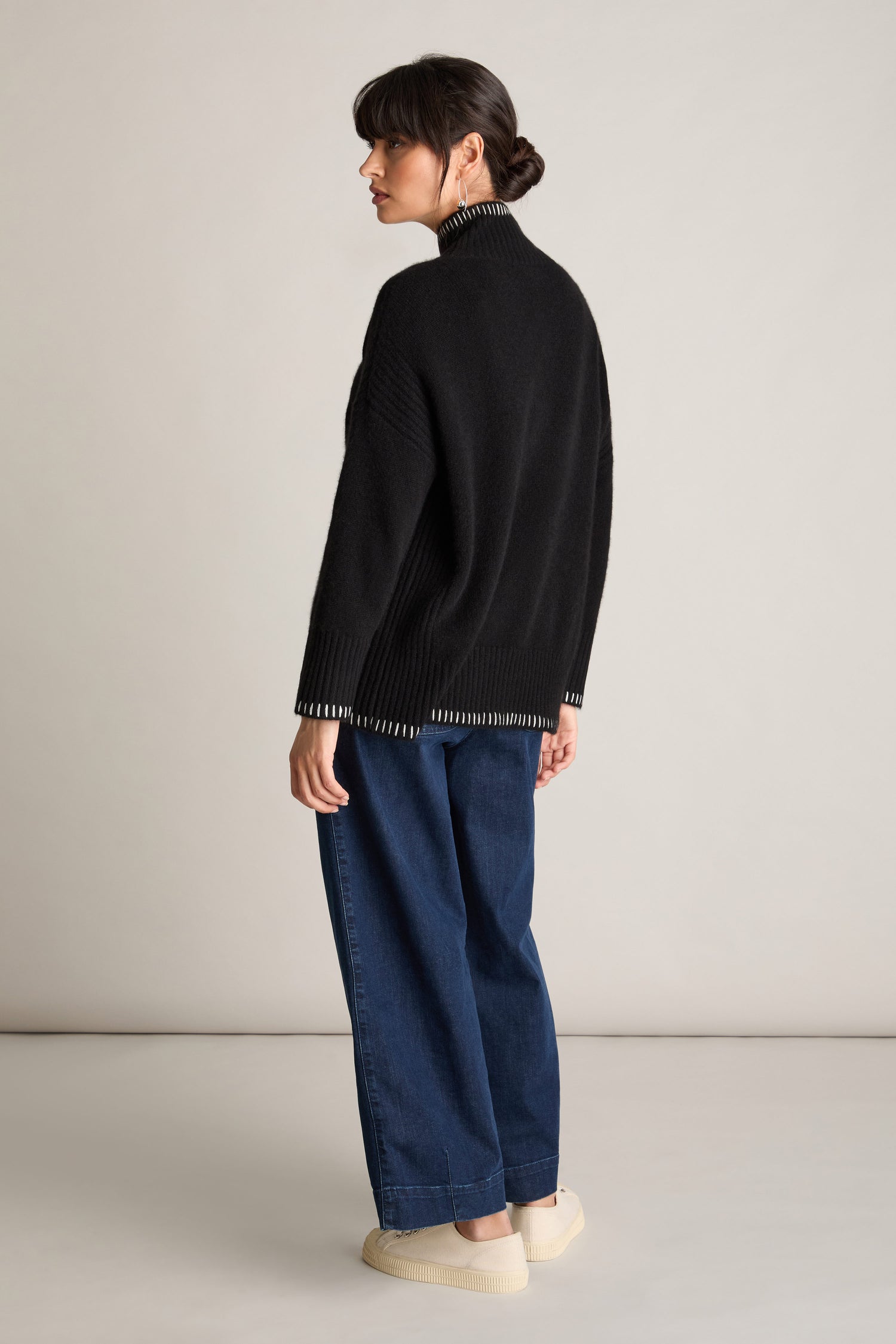 A woman stands with her back to the camera, wearing a black Blanket Stitch Funnel Neck Cashmere Knit sweater, wide-leg denim pants, and white shoes. Her dark hair is styled in a low bun.