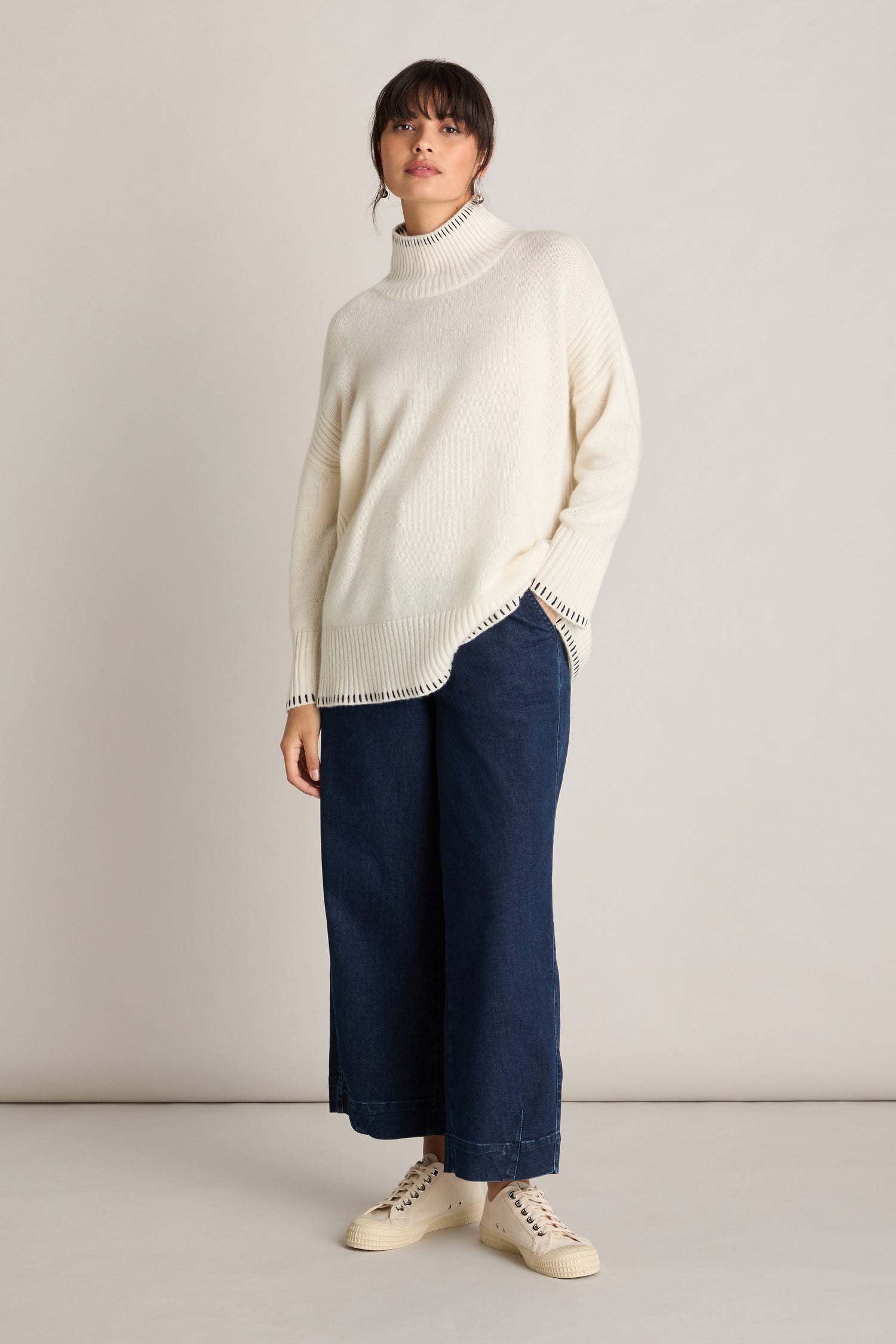 A person stands against a plain background wearing an Estheme Blanket Stitch Funnel Neck Cashmere Knit in cream, dark wide-leg trousers, and white sneakers.