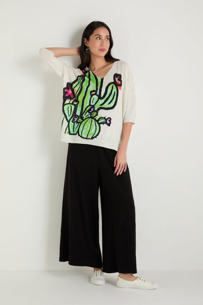 A woman stands against a white wall wearing the Cactus Linen Cotton V-Neck Knit, featuring a colorful cactus design, paired with black wide-leg pants and white sneakers.