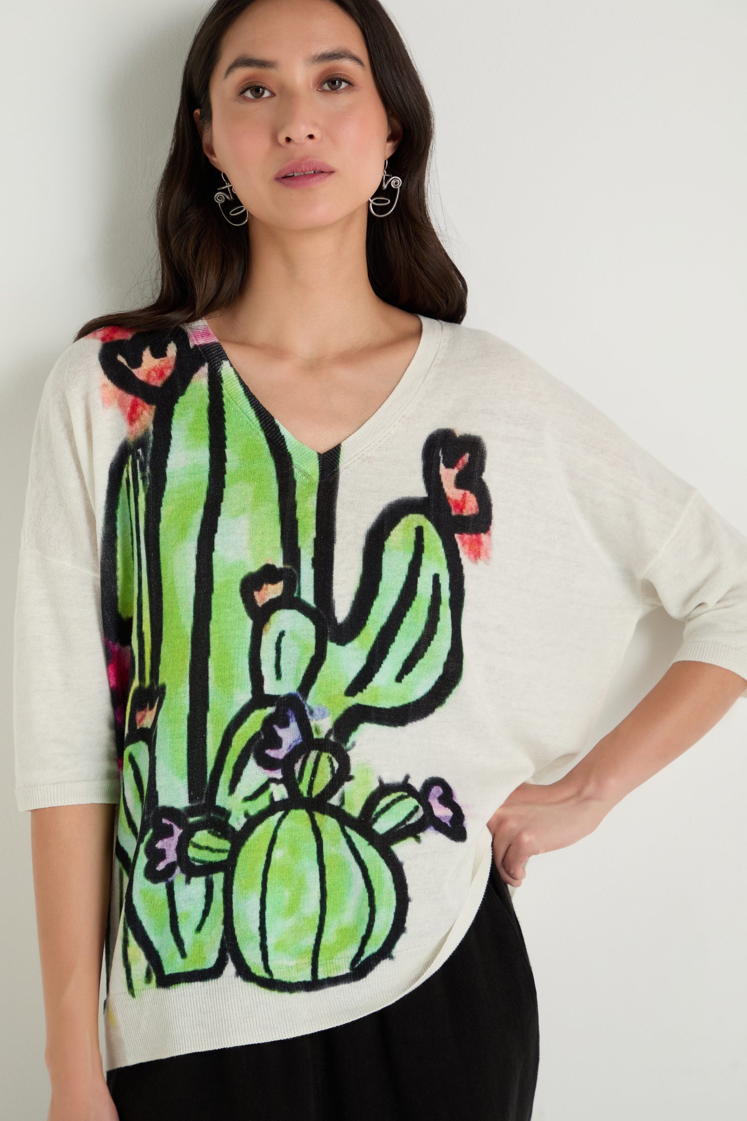 A woman models the Cactus Linen Cotton V-Neck Knit, a white sweater with a colorful cactus and flower graphic, standing against a plain light background with one hand on her hip.