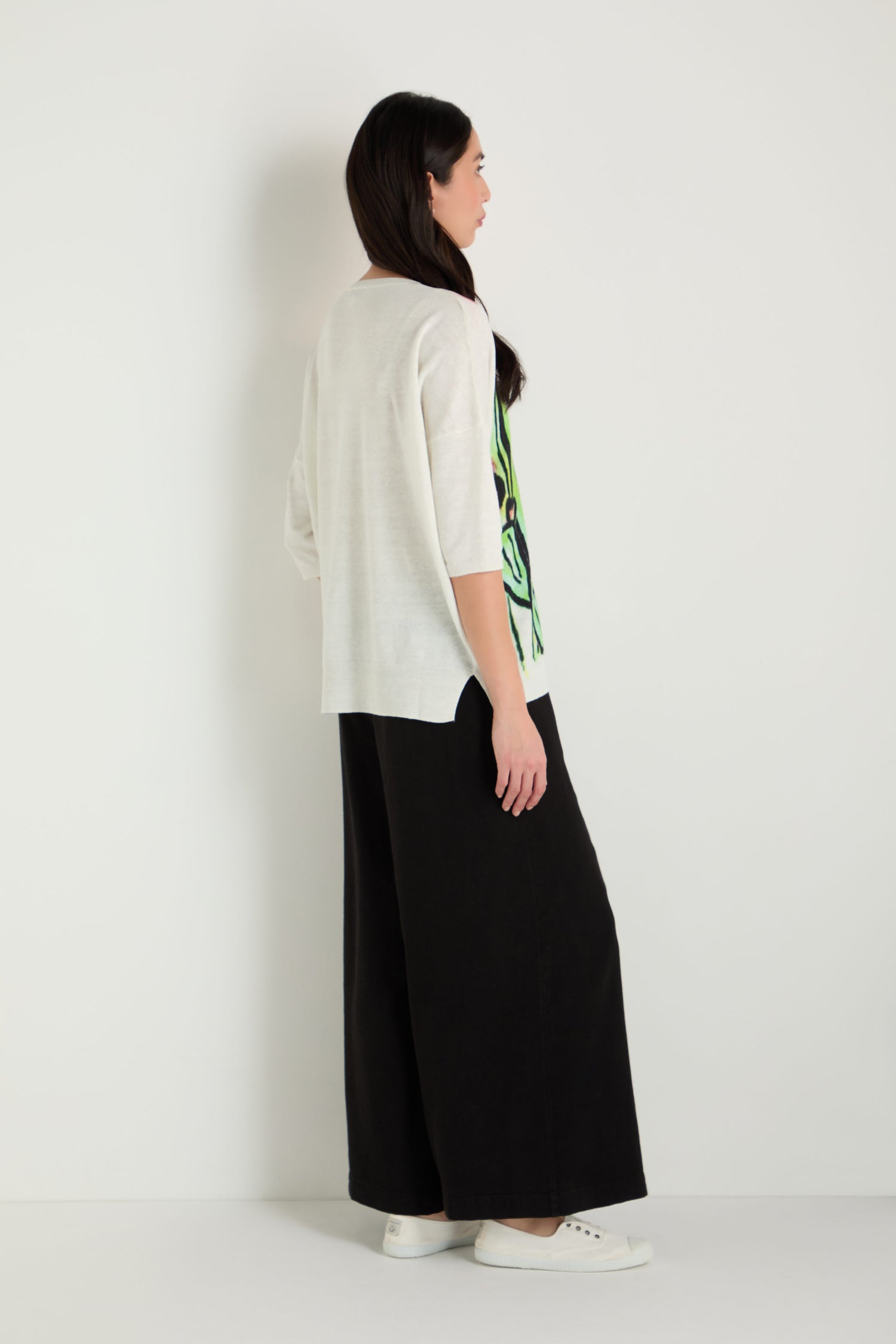 A woman stands sideways against a white background, wearing the Cactus Linen Cotton V-Neck Knit top with wide-leg black pants and white sneakers.