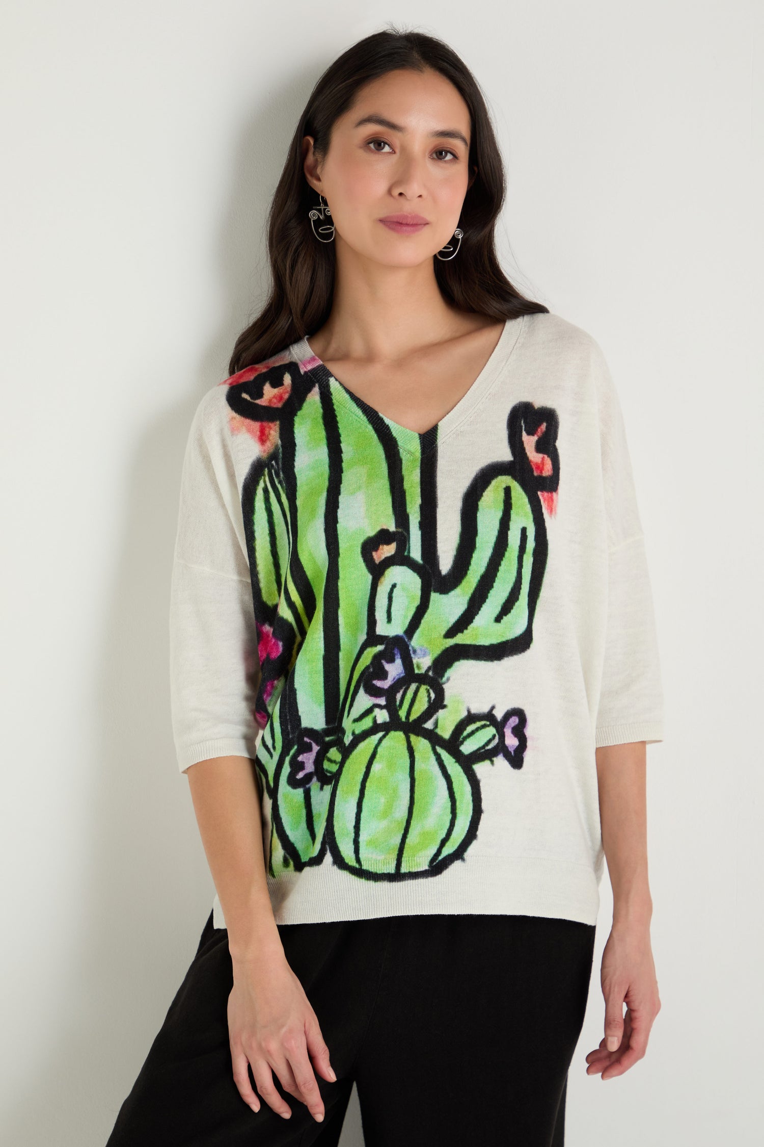 A woman stands against a white wall wearing the Cactus Linen Cotton V-Neck Knit, a white sweater with a large colorful cactus graphic, paired with black pants.