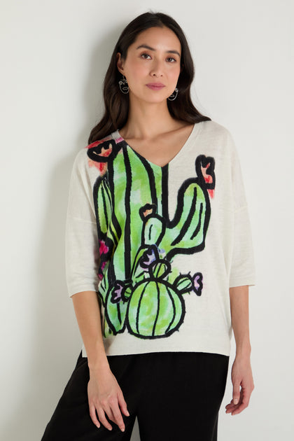A woman stands against a white wall wearing the Cactus Linen Cotton V-Neck Knit, a white sweater with a large colorful cactus graphic, paired with black pants.