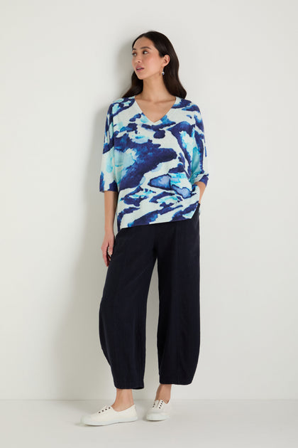 A woman stands against a white wall wearing the Cloudy Skies Linen Cotton V-Neck Knit, loose black pants, and white sneakers.