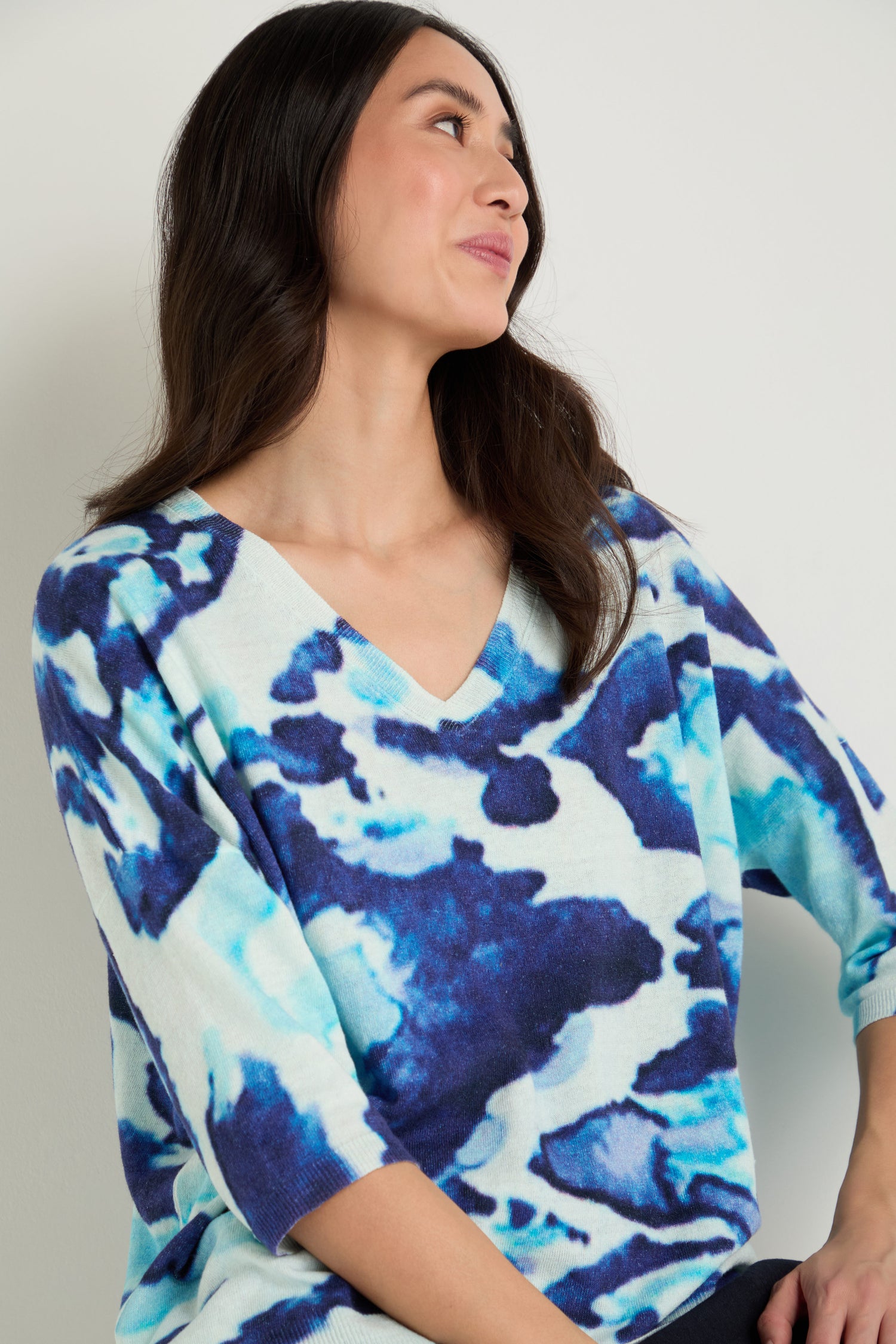 Woman with long dark hair wears the Cloudy Skies Linen Cotton V-Neck Knit, featuring a blue and white abstract cloudy print, and looks to the side against a plain background.