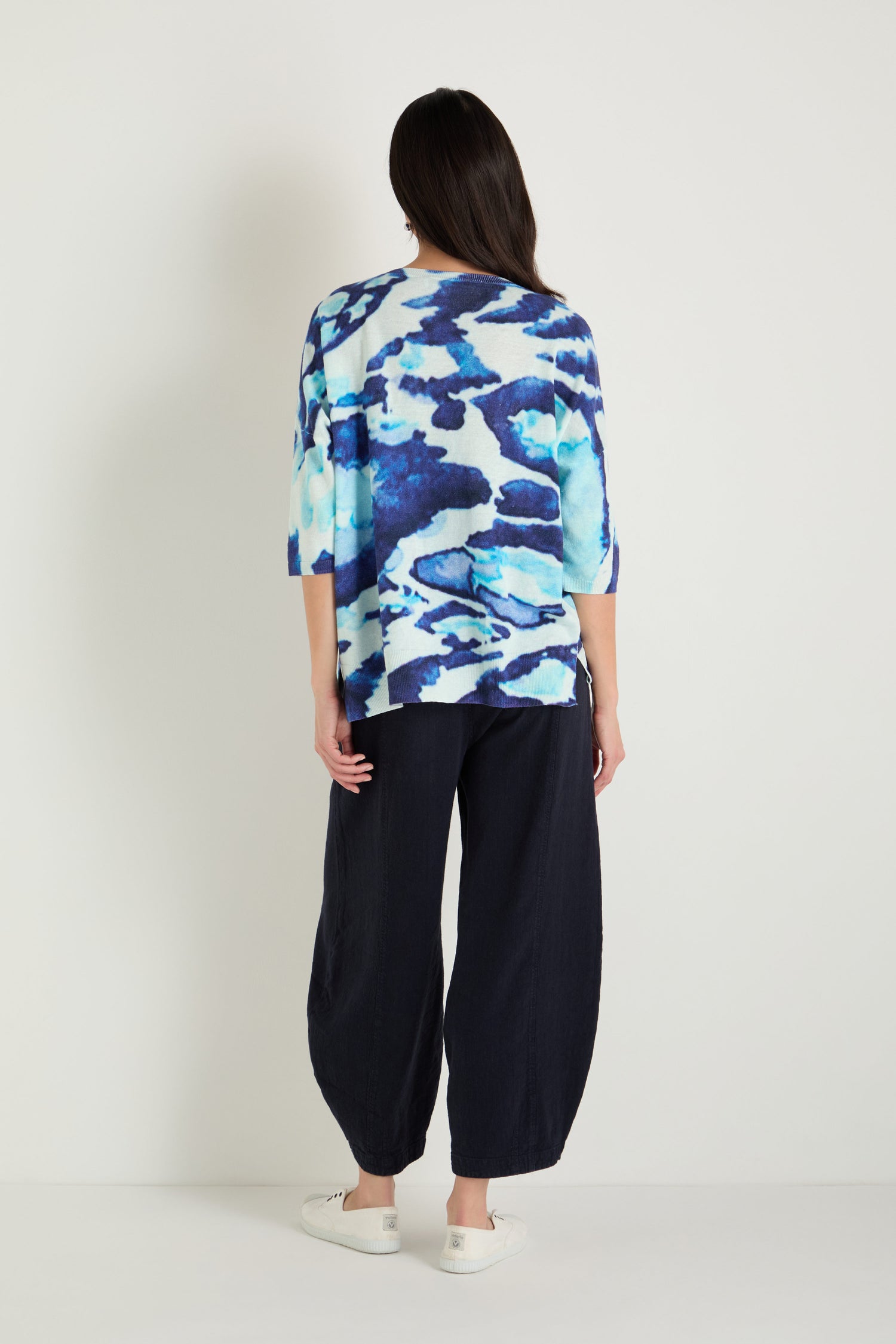 A person faces away, wearing the Cloudy Skies Linen Cotton V-Neck Knit with an abstract print, loose dark navy pants, and white slip-on shoes against a plain light background.