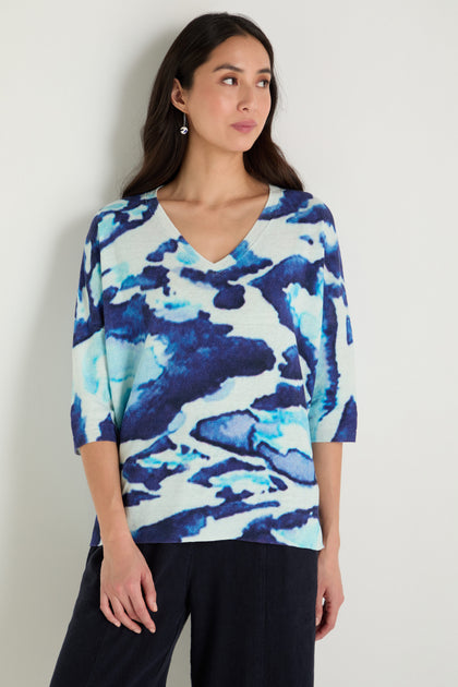 Woman wears the Cloudy Skies Linen Cotton V-Neck Knit, featuring an abstract blue and white cloudy print, paired with dark pants and standing against a plain light wall.