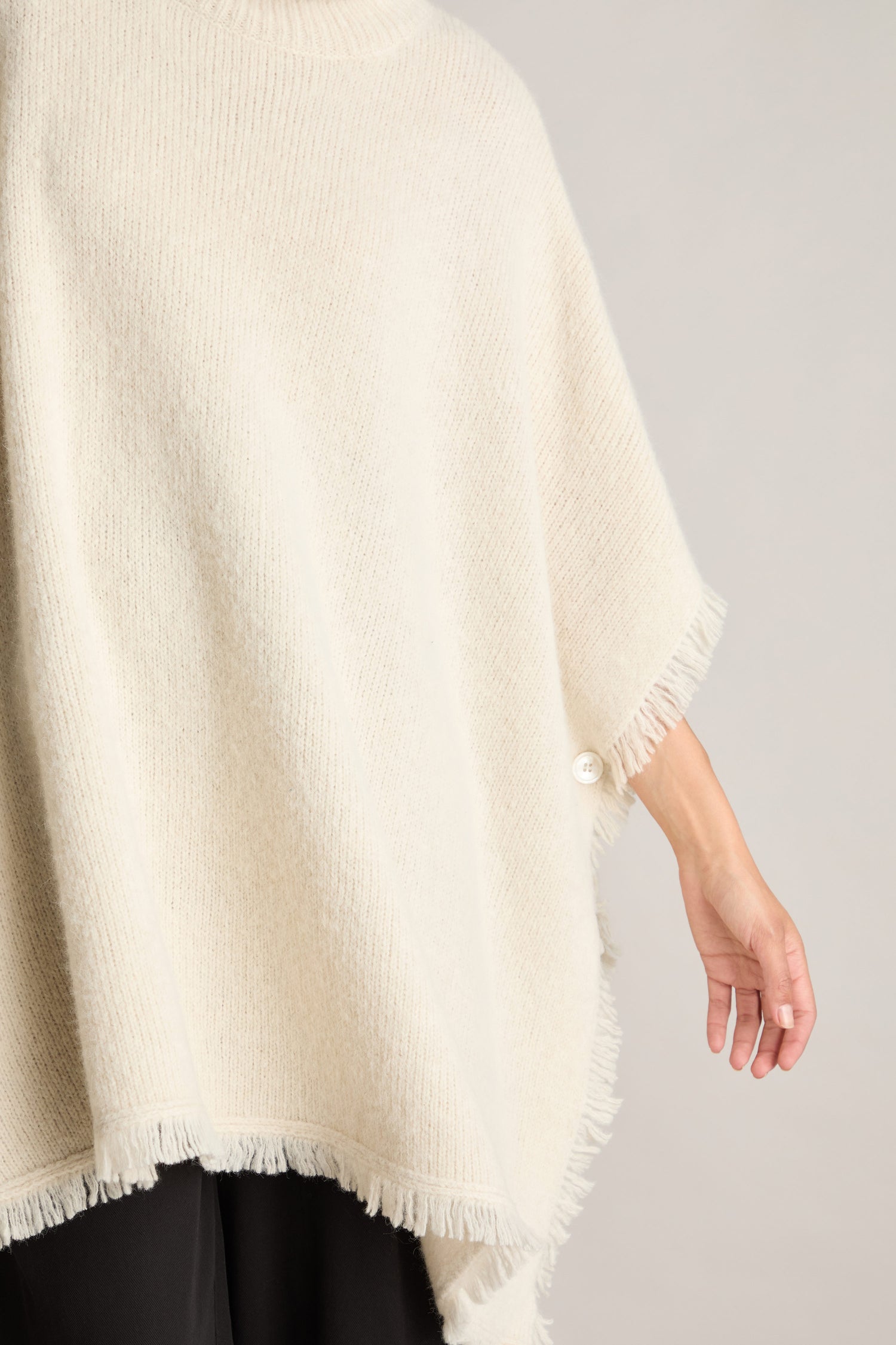 Close-up of a person wearing the Cashmere and Alpaca Poncho in cream, featuring fringe detailing, a polo neck, and an elegant button on the side. The person is shown from the back and side with their arm extended outward, exuding an aura of luxury and comfort.