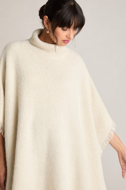 A person wearing the Cashmere And Alpaca Poncho, featuring a white, high-neck, and knitted design, looks down to their left side, exuding luxury and comfort.