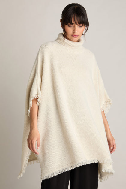 A person wearing a loose, off-white Cashmere And Alpaca Poncho with fringed edges stands against a plain background, epitomizing luxury and comfort.