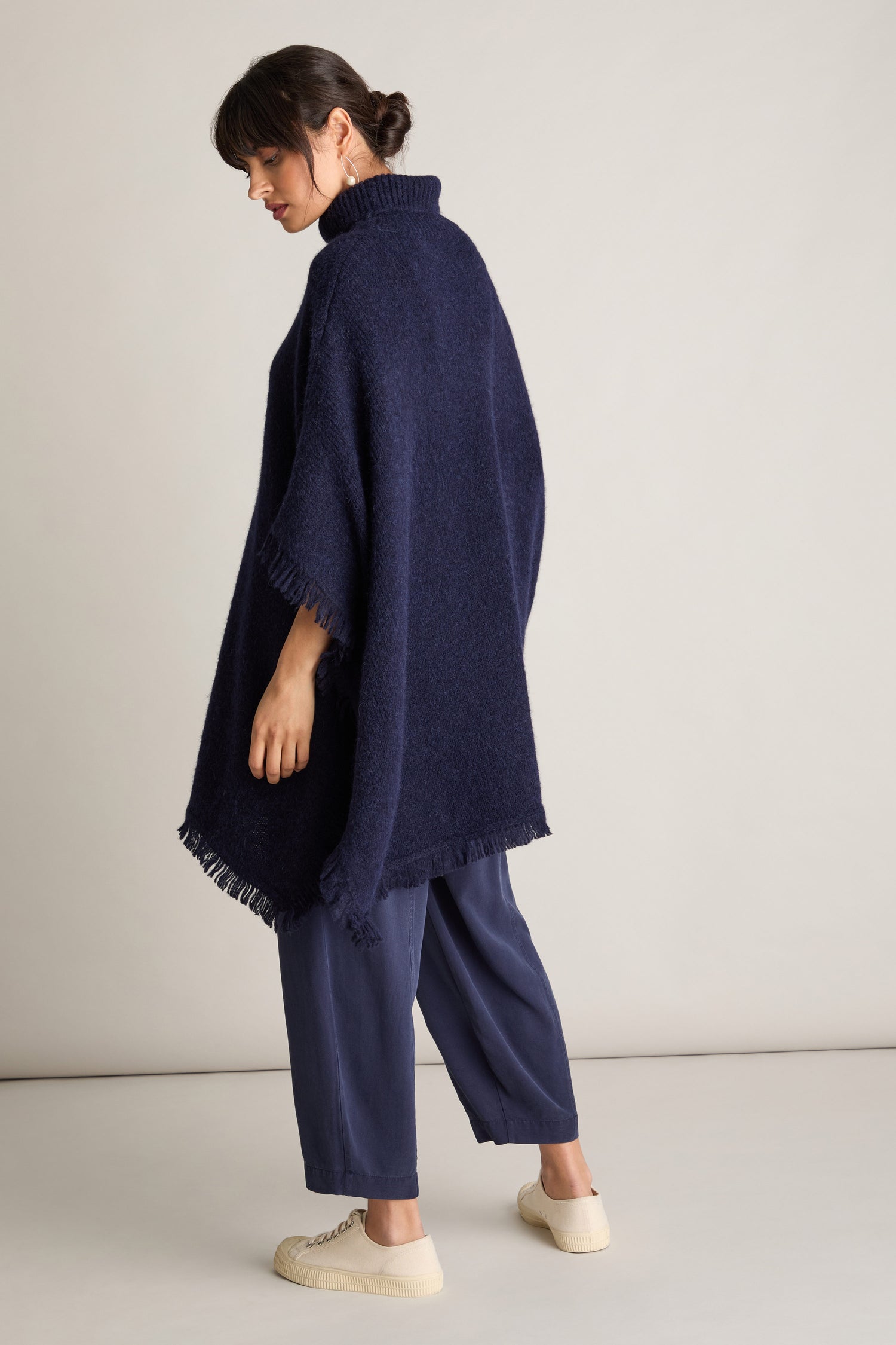 A woman standing sideways, with her hair tied back, dons a luxurious dark blue Cashmere And Alpaca Poncho over loose blue pants and white shoes, radiating elegance and comfort.